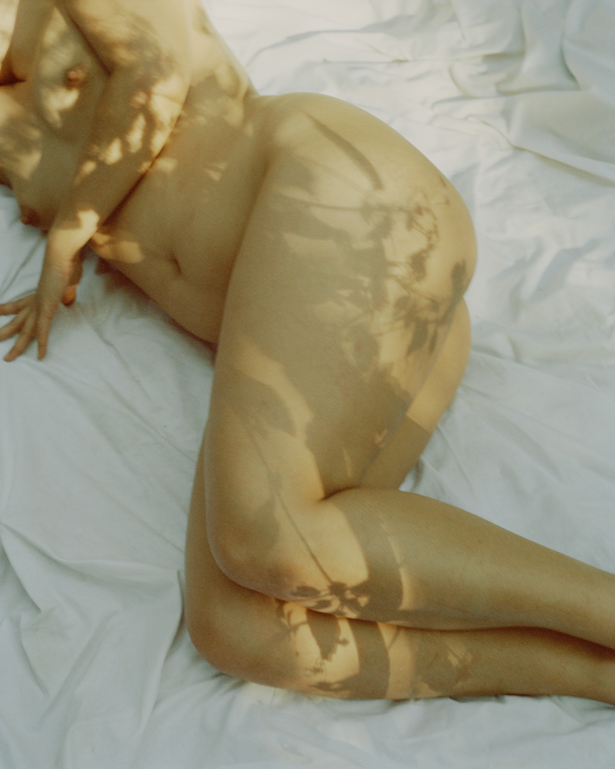Colour photograph depicting girl lying on white cloth with dappled sun on bare skin.
