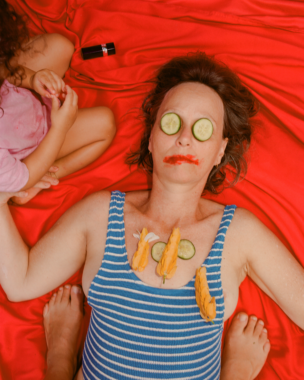 Colour photograph depicting woman lying on red cloth with cucumbers on eyes and smeared red lipstick on her mouth. 