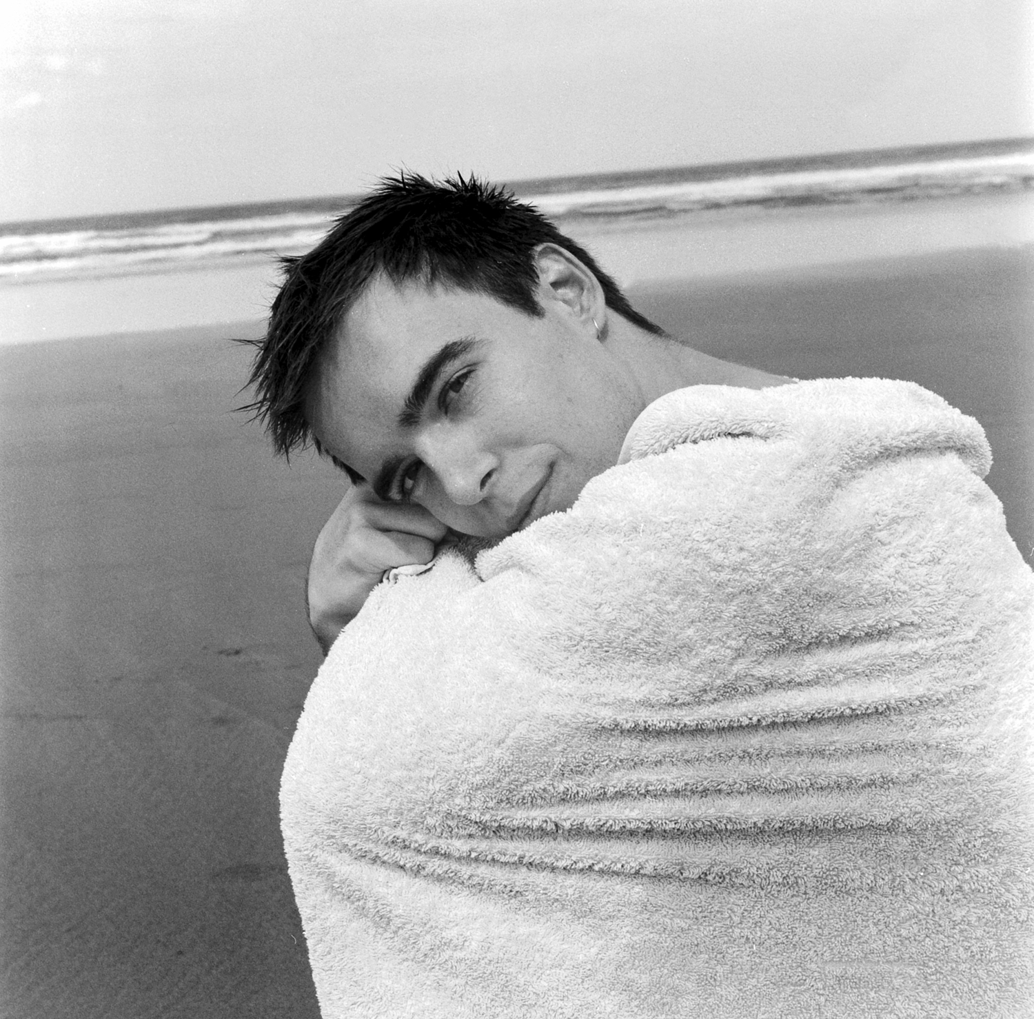 Black and white photograph depicting a portrait of a man wrapped in towel sitting on beach.
