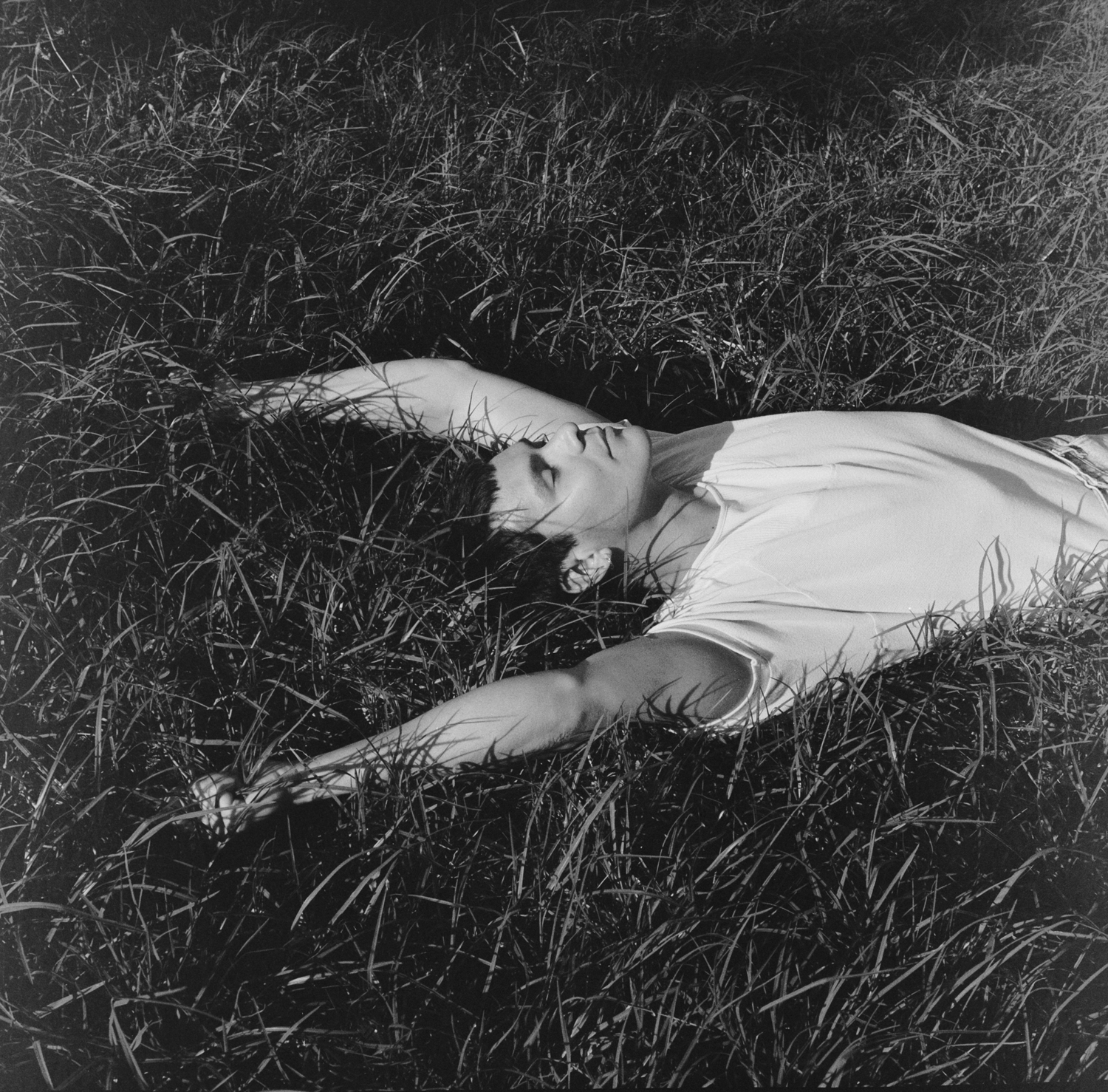 Black and white photograph of a man in a white shirt asleep in a grass field.