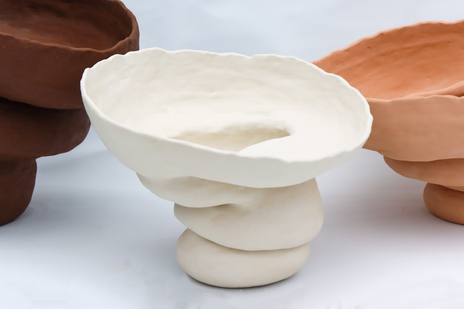 Close up image of three large voluptuous ceramic forms lined up in front of a white backdrop, coloured from left to right, dark brown, white and pink.