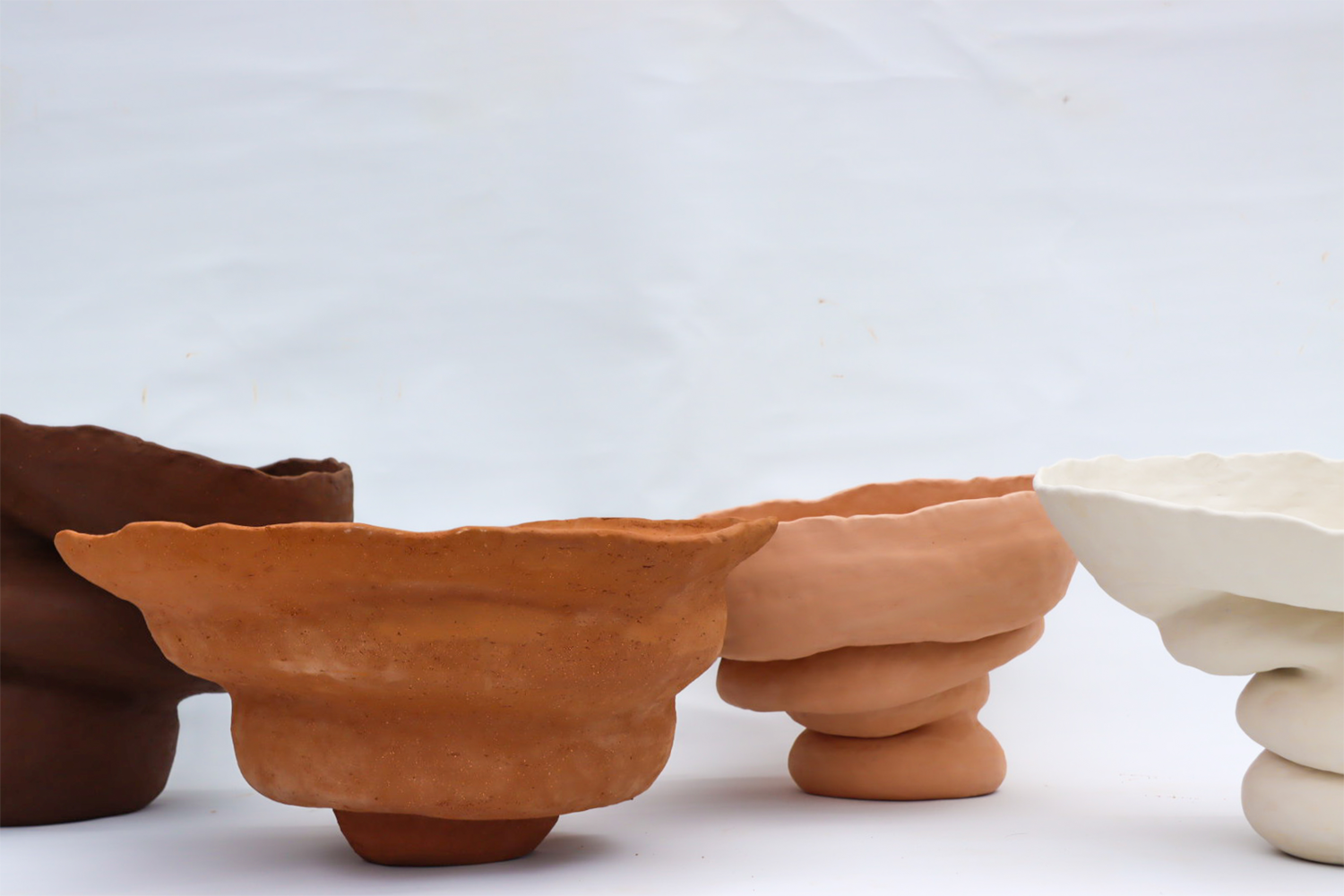 Four large voluptuous ceramic forms lined up in front of a white backdrop, coloured from left to right, dark brown, mid toned orange, pink and white.