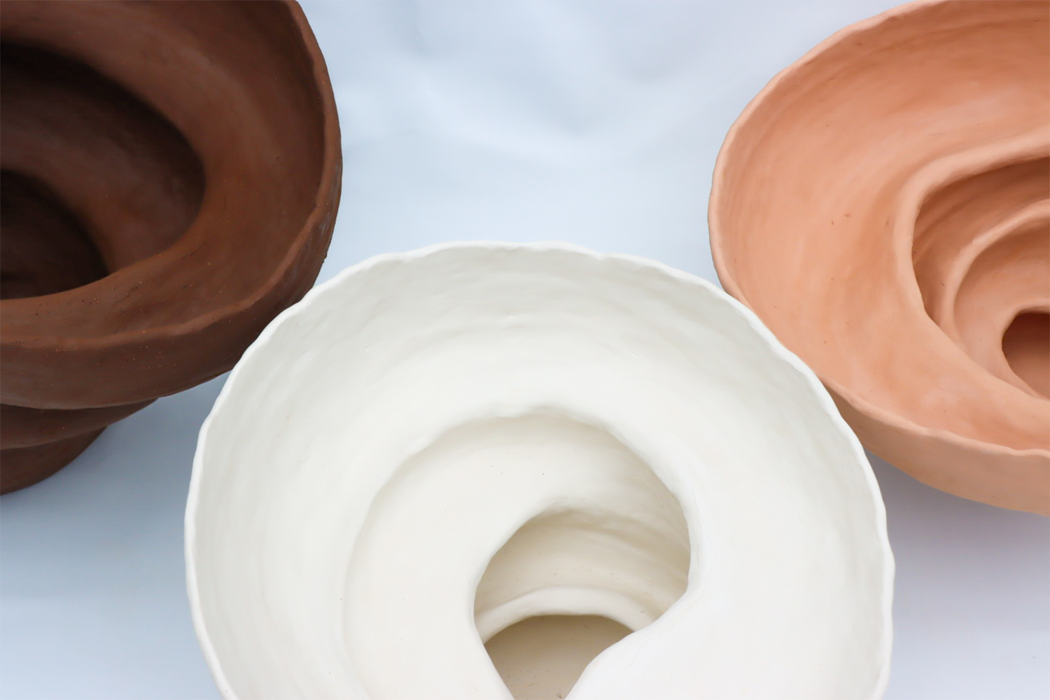 Three large voluptuous ceramic forms in a top down view atop a white backdrop, coloured from left to right, dark brown, white and pink.
