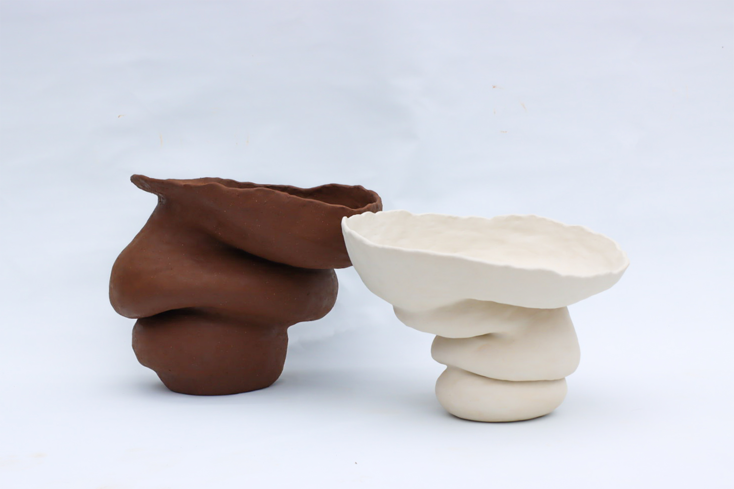 Two large voluptuous ceramic forms in front of a white backdrop, coloured from left to right, dark brown and white.