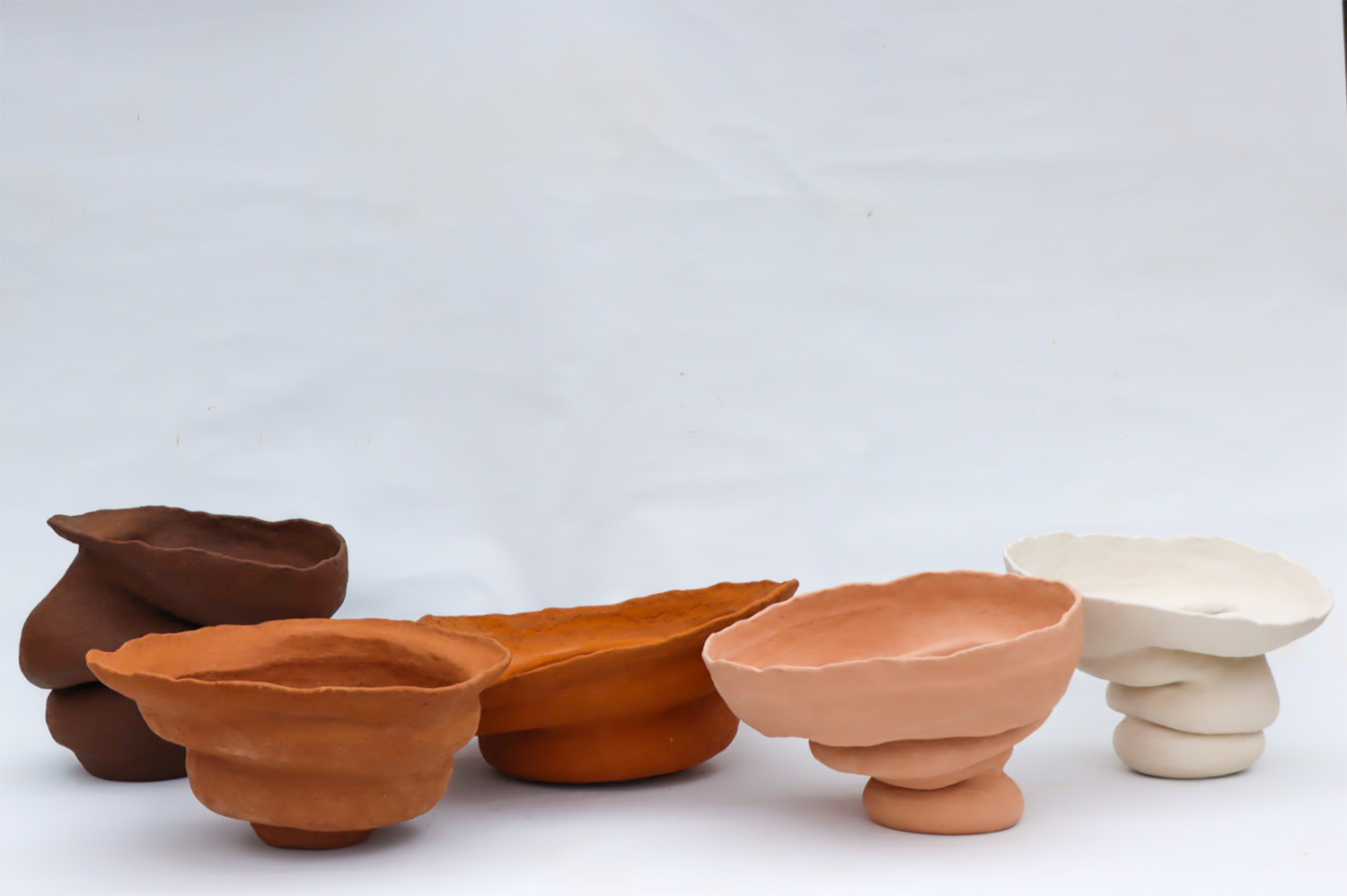 Five large voluptuous ceramic forms in a line in front of a white backdrop, coloured from left to right, dark brown, mid toned orange, dark toned orange, pink and white.