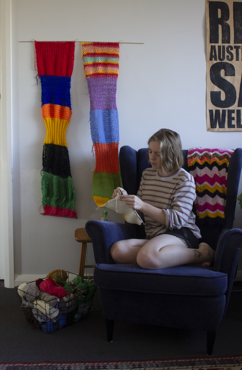 A woman sits in an armchair knitting. The knitting is hanging on the wall while she knits.