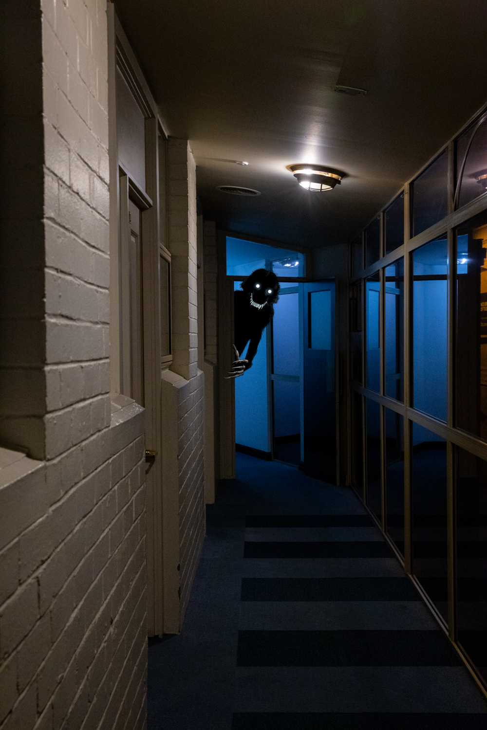 Colour photograph depicting a hotel corridor with a dark creature peering out from behind a door.