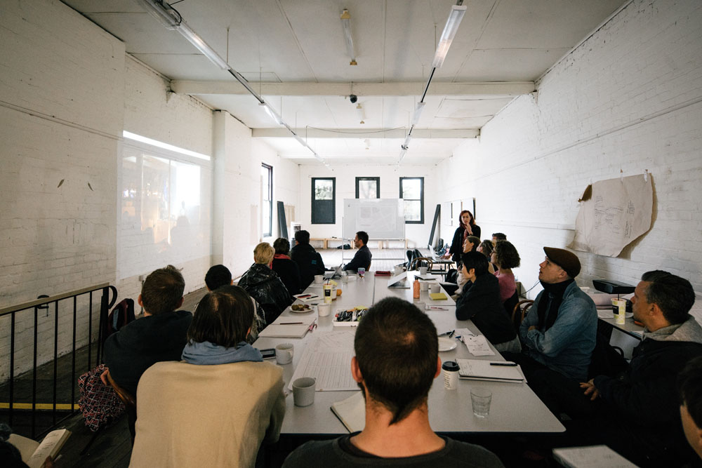 Artists sitting around a table, viewing a presentation by Natalie King.