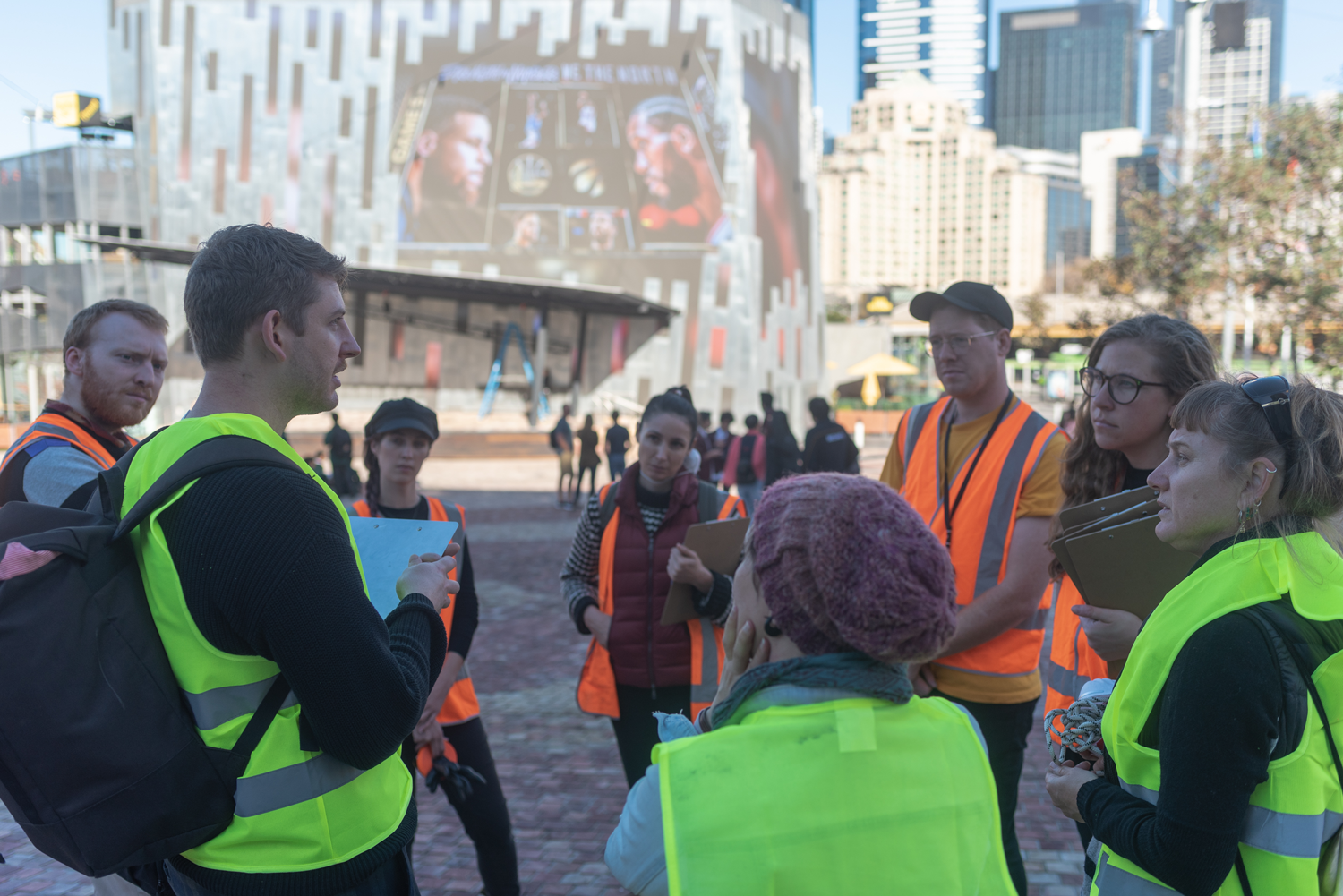 Artists stand in a circle at Fed Square. They are all wearing hi-vis vests.
