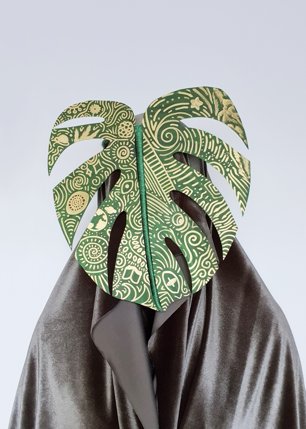 A green cloaked figure stands against a white background. They wear a green paper leaf with splits over their head. The leaf is covered with golden designs, some swirl whilst some resemble objects. 