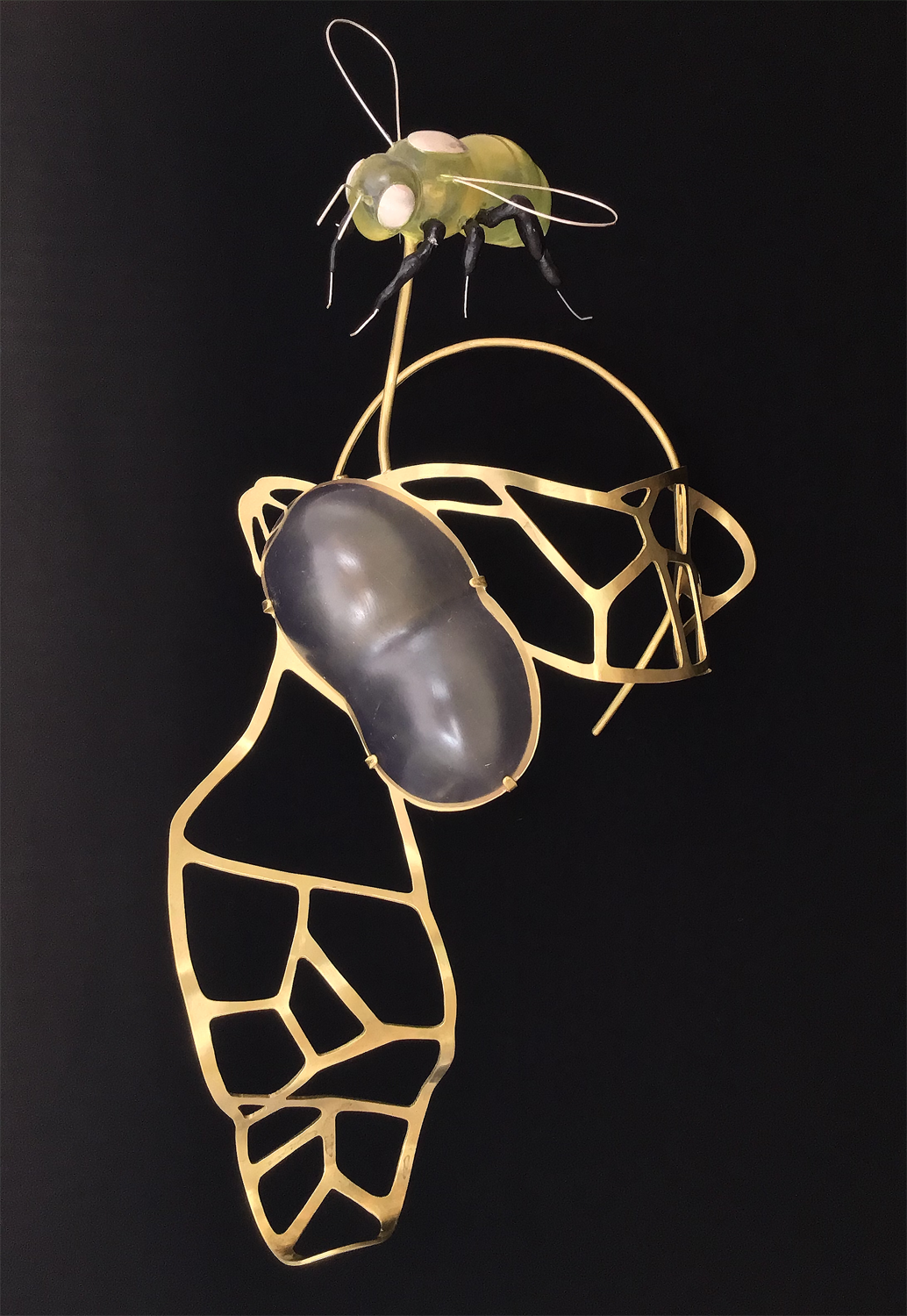 A queen bee hovers over an abstract brass form. The brass form resembles wings; they both join a translucent and elongated eye-like form. 