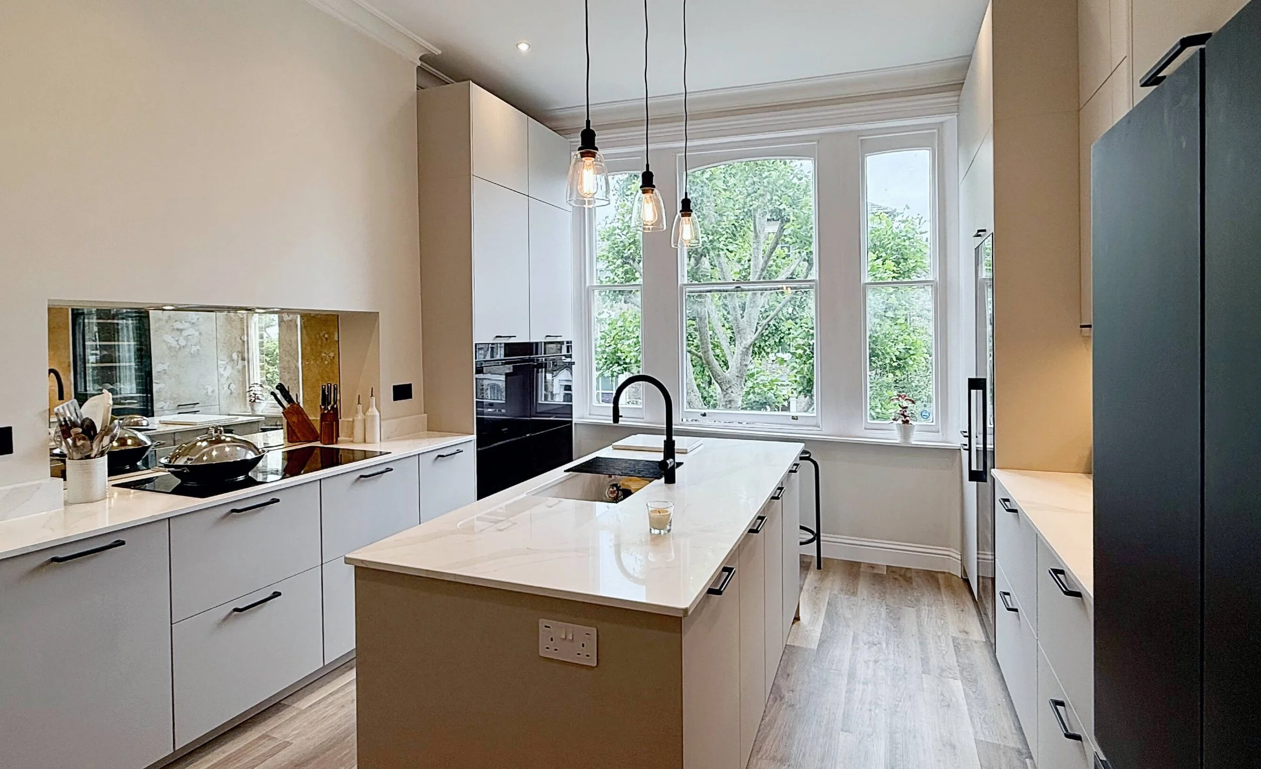 Bright modern kitchen with white cabinets, large island, black fixtures, pendant lighting, and mirrored backsplash under wide windows.