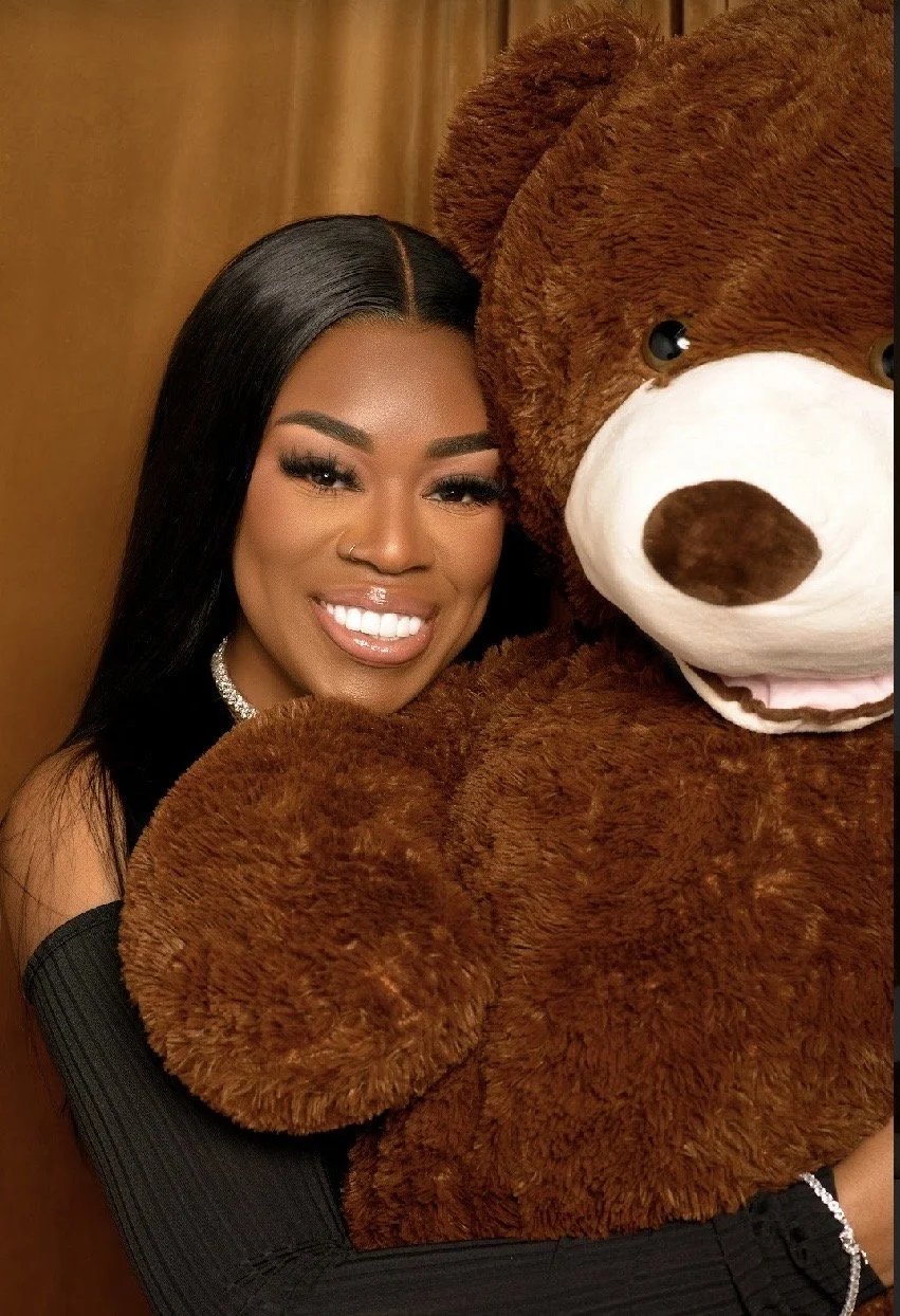 A woman with long black hair, smiling, holding a large brown teddy bear with a white snout and dark nose.