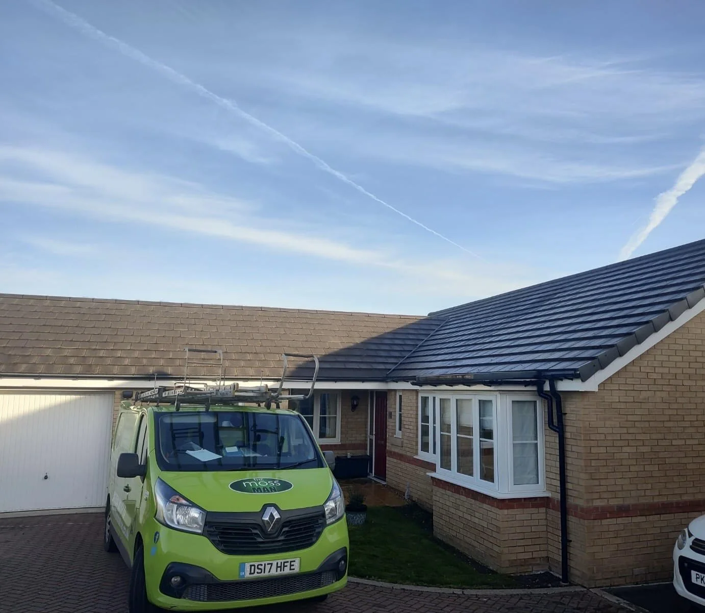 A house with brick exterior, white window frames, and a brown tiled roof. A bright green Moss Man service van with a ladder on top is parked in the driveway. The sky above is blue with a few contrails.
