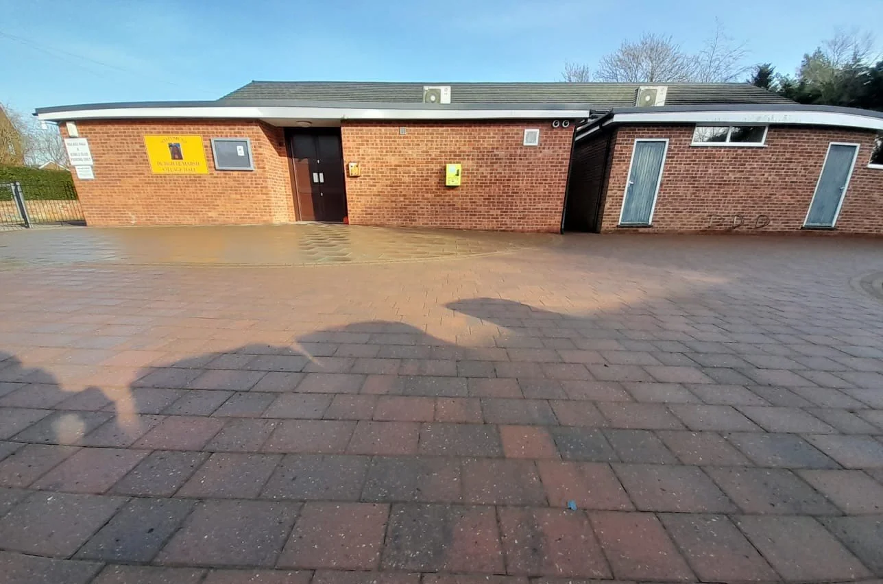 A brick building with two doors and a yellow sign on the left, surrounded by a brick-paved area. Shadows of flags are cast on the ground in the foreground.