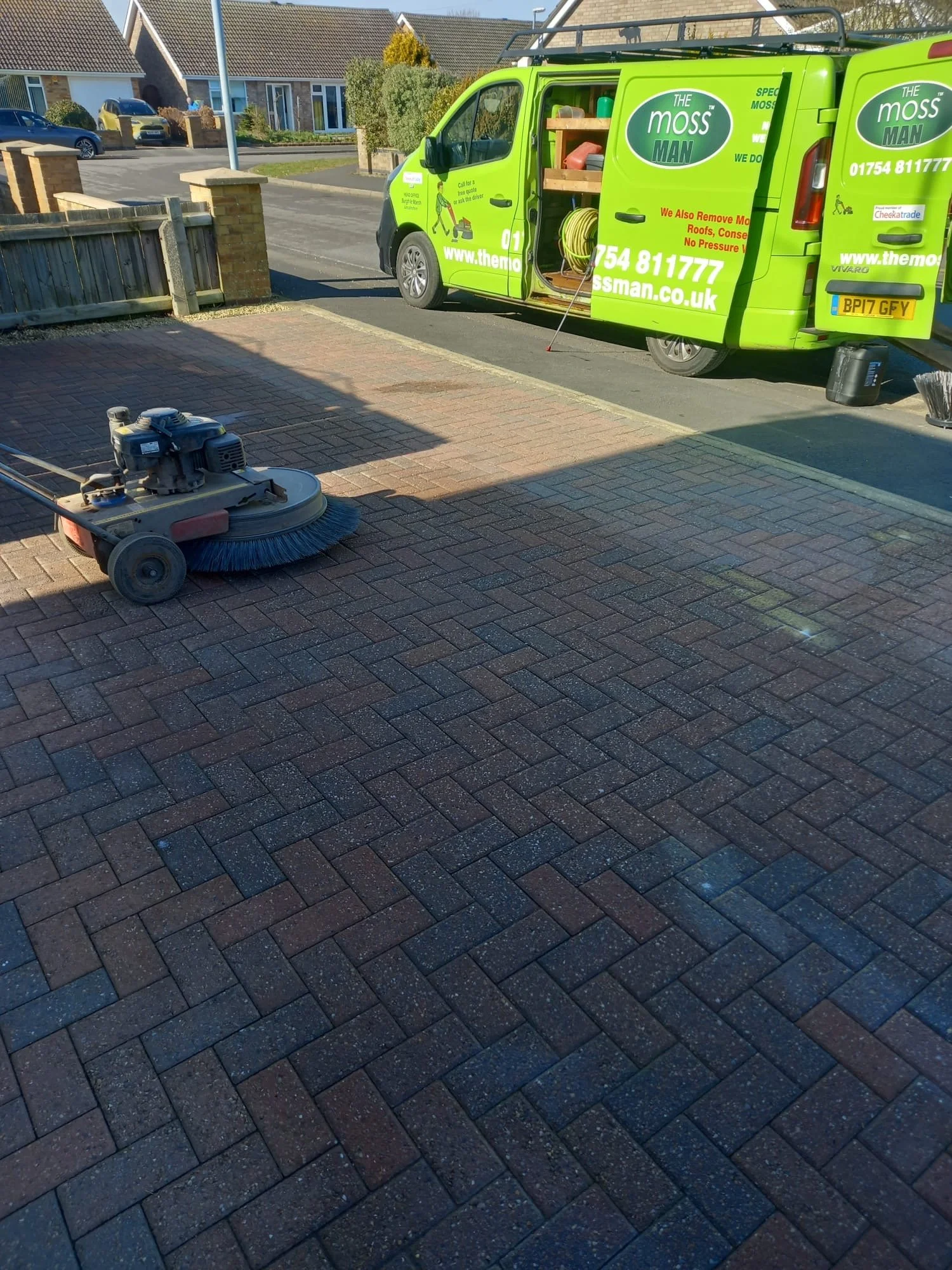 A road sweeper machine on a paved driveway, with a green moss removal service van parked on the street in the background.