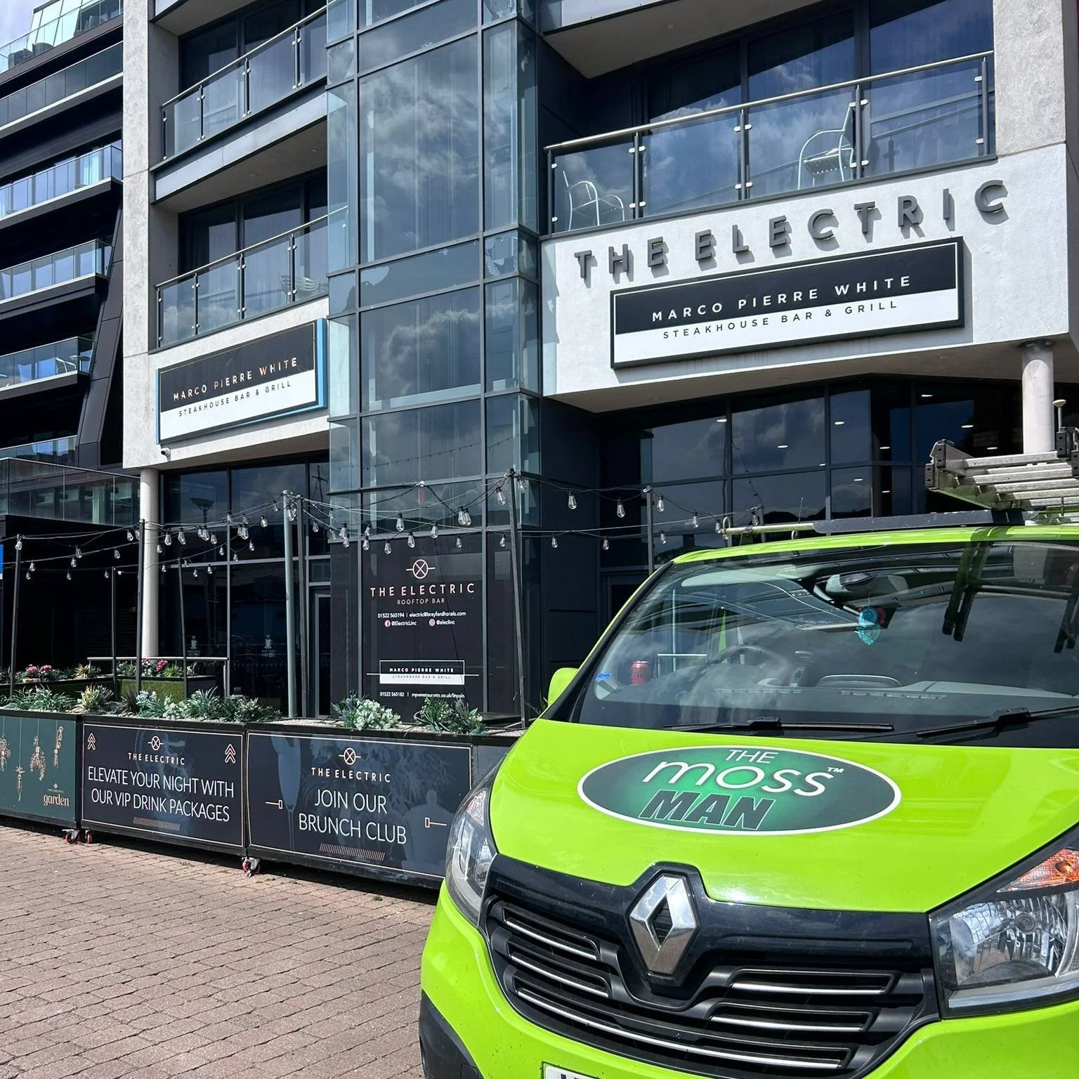 Exterior view of a modern building with glass balconies and signage for Marco Pierre White Steakhouse Bar & Grill and The Electric Rooftop Bar. A bright green van, labeled 'The Moss Man,' is parked in front.