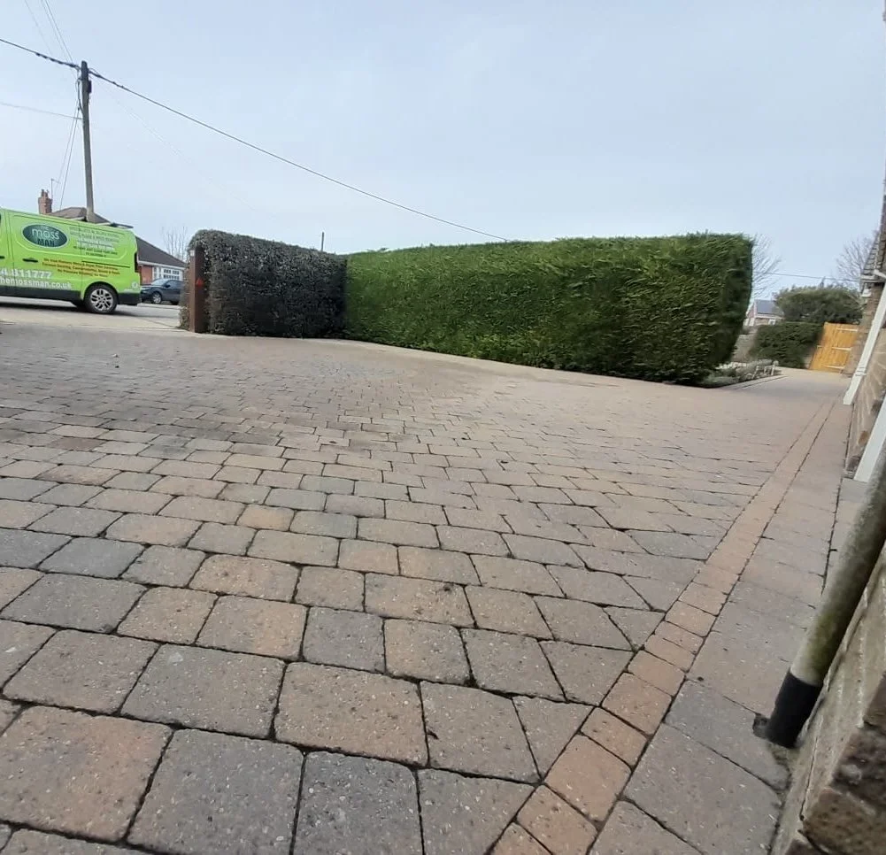 A paved driveway with red and gray bricks, bordered by a stone wall on the right and trimmed hedges in the background, with a green maintenance vehicle parked on the street.