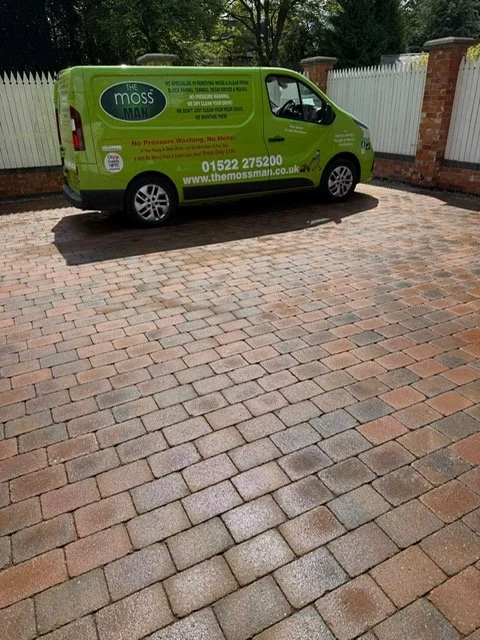 Green Moss Man service van parked on a brick driveway with a white fence and trees in the background.