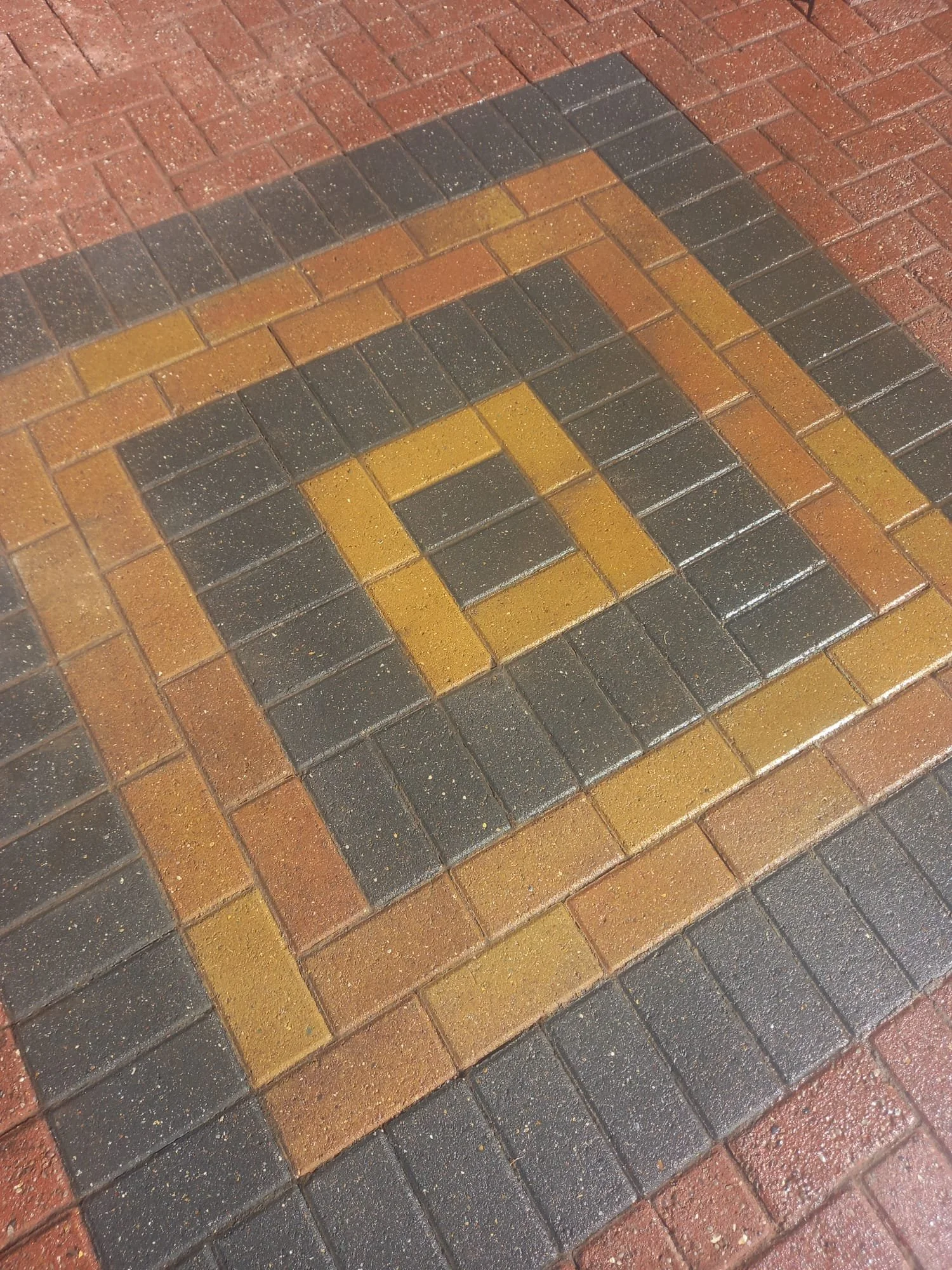 Pattern of brick pavement with a central square design made of yellow and black bricks, surrounded by brown and red bricks.