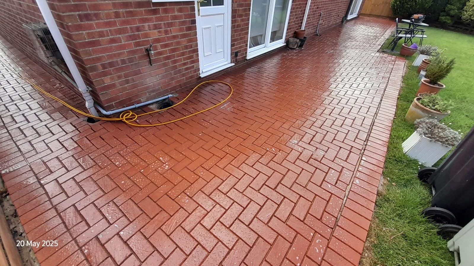 Wet red brick patio with garden tools, potted plants, and a black trash bin near a brick house with sliding glass doors and white door, surrounded by a lawn and garden.
