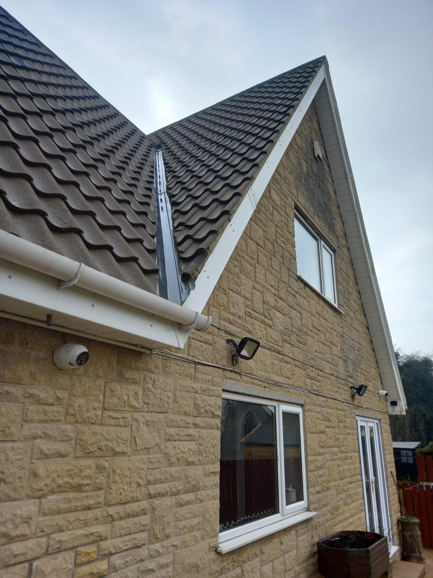 The side of a house with beige brick walls, a window, a glass door, outdoor security cameras, and exterior lights. The roof has a steep pitch with dark brown tiles and includes a rain gutter and a metal downspout.