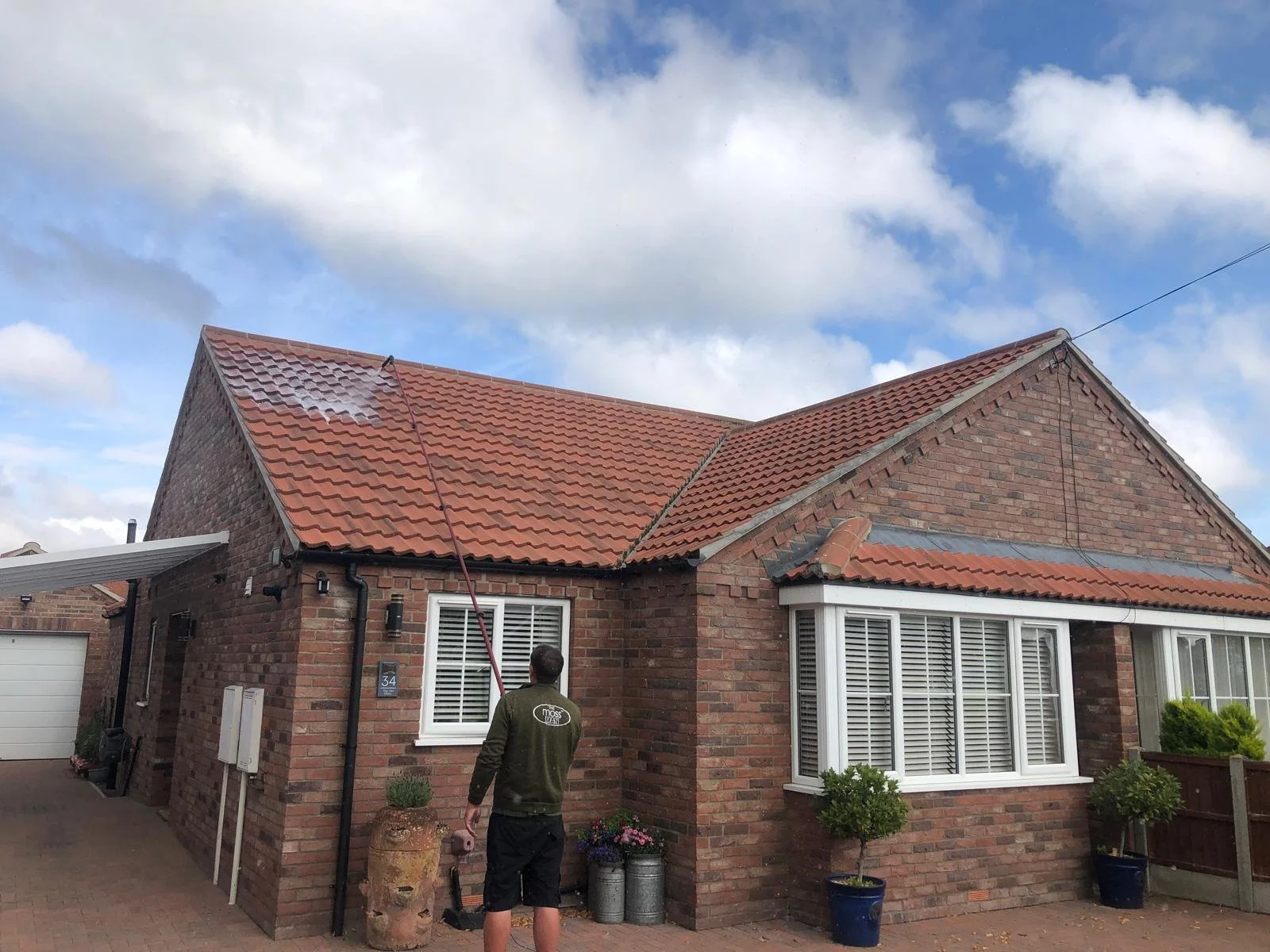 A man on a ladder cleaning the roof of a brick house with a pressure washer. The house has a red tiled roof, a bay window, and potted plants outside. The sky is partly cloudy.