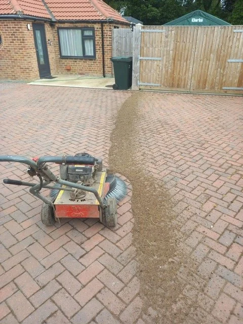 A lawn care machine is blowing sand or gravel across a brick driveway, creating a narrow streak of accumulated material leading towards a wooden fence.