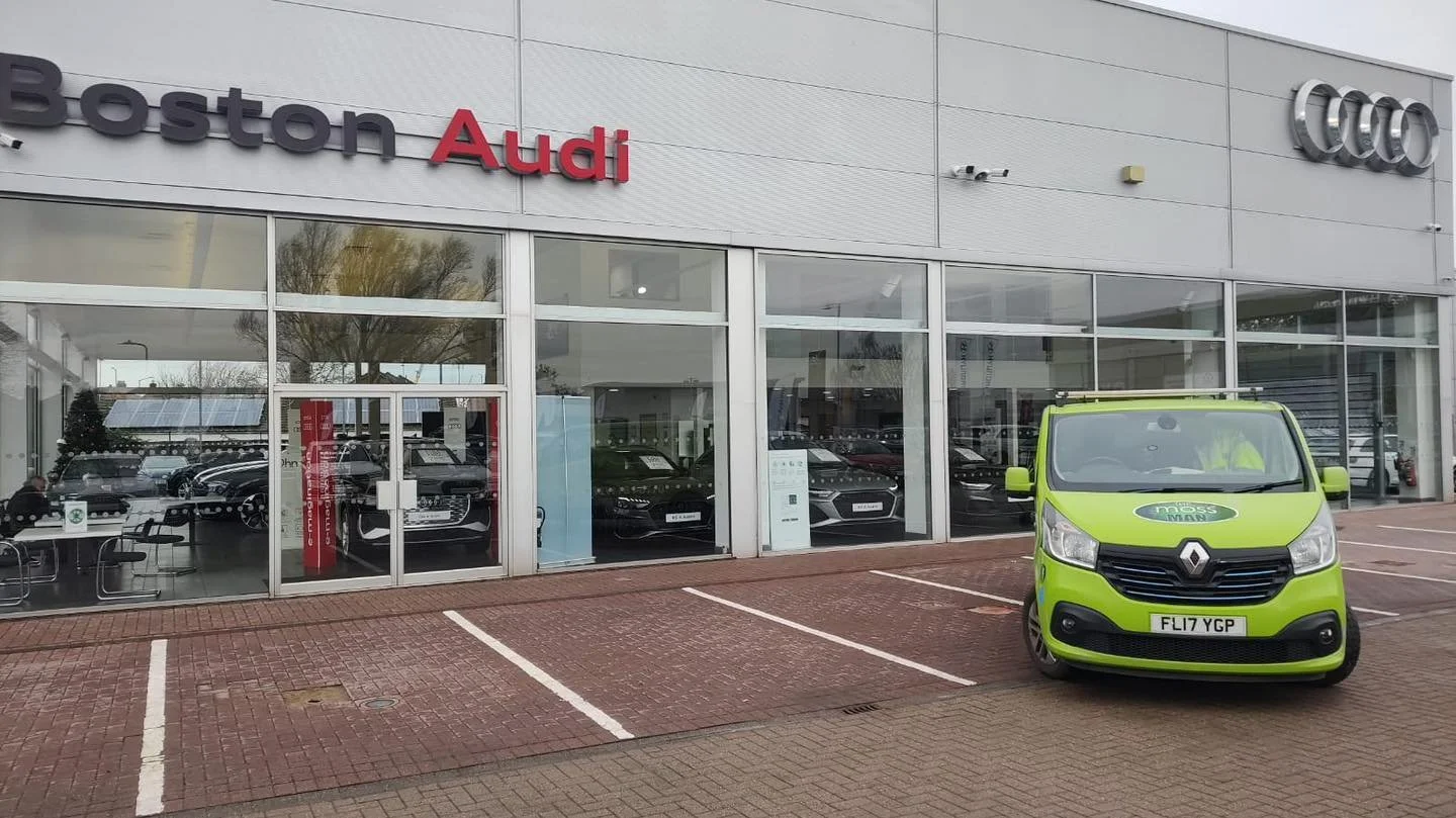 Exterior of a car dealership with the signs 'Boston Audi' and four interlinked silver rings logo on the building. A green Renault vehicle is parked in front.