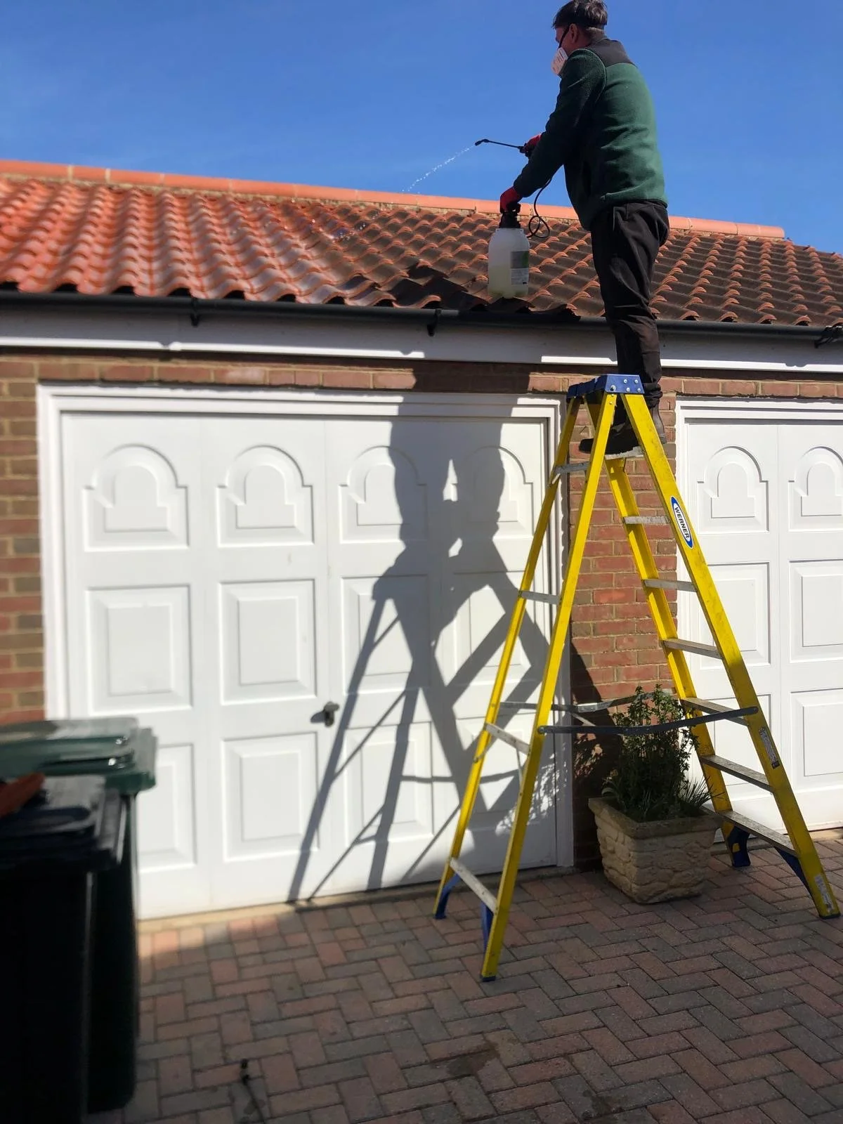 Person standing on a yellow ladder cleaning a tiled roof with a spray nozzle, with a clear blue sky in the background.