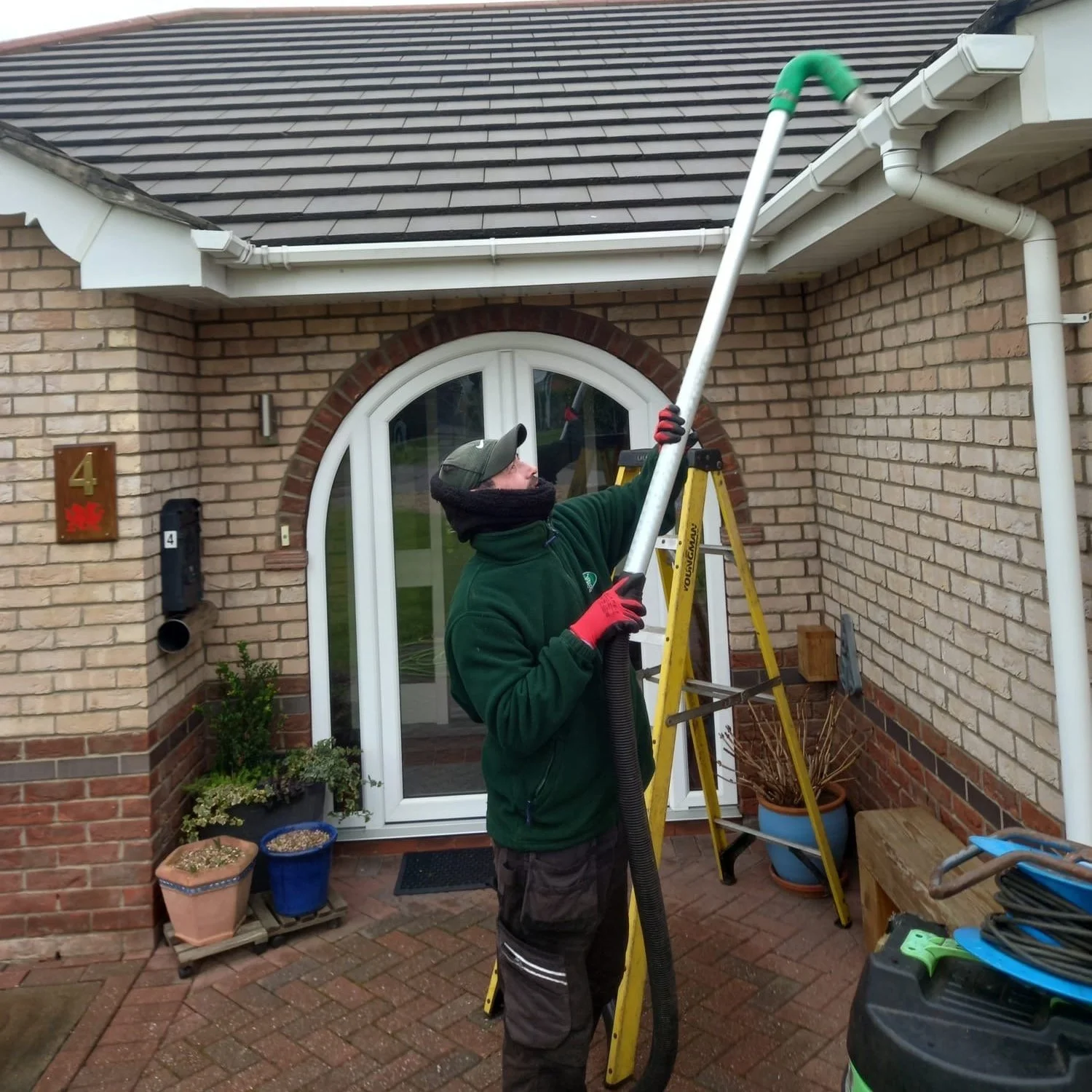 A man wearing a green jacket, red gloves, and a cap is cleaning the high gutter of a brick house using a long pole attached to a vacuum hose. He is standing on a yellow step ladder in the front yard, which has potted plants and garden tools.