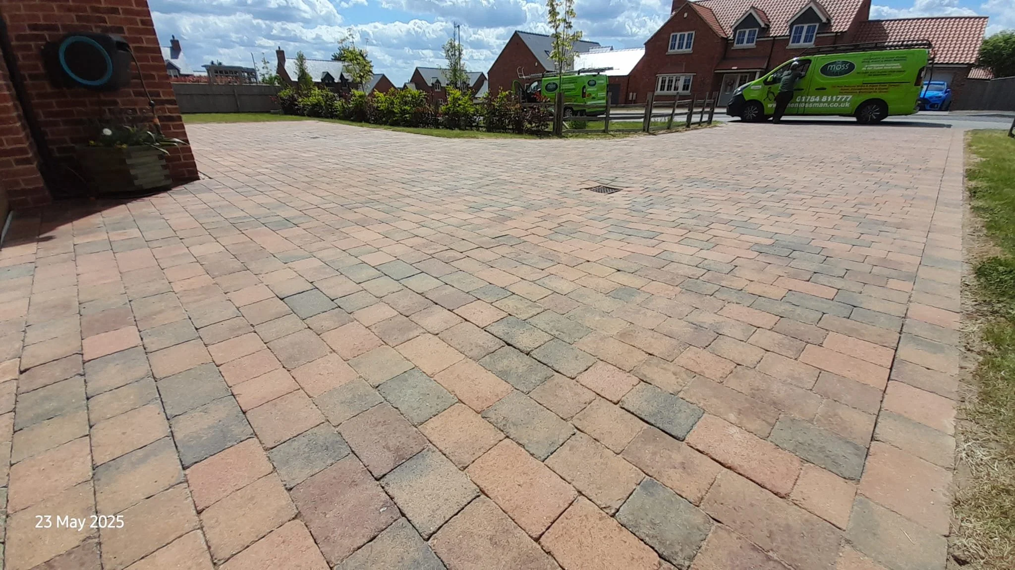 A newly paved driveway with colored bricks, a garden with shrubs and young trees, two green moss service vans parked near a residential street, and a person near one of the vans. The sky is partly cloudy.