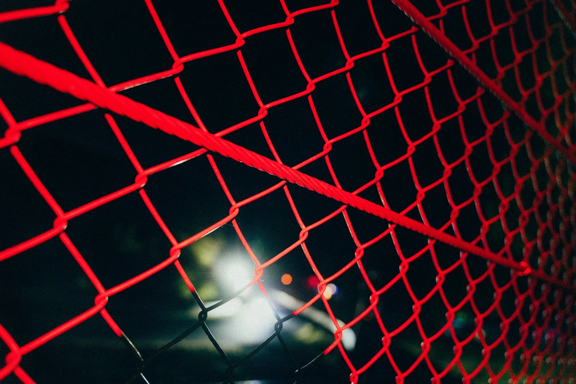 Close-up of a red and black chain-link fence at night, with blurred city lights in the background.
