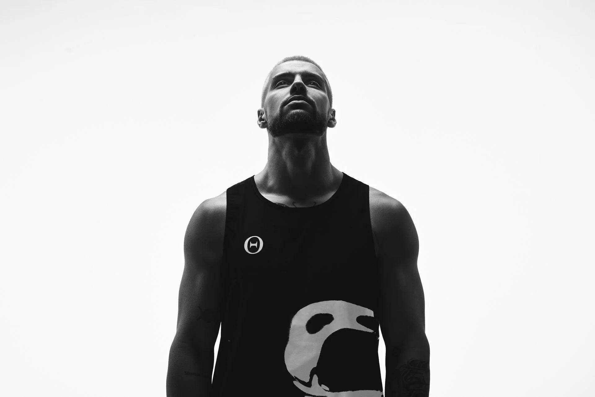 A black and white photo of a man with a beard, wearing a tank top with a skull graphic, looking upwards with a serious expression, against a plain light background.