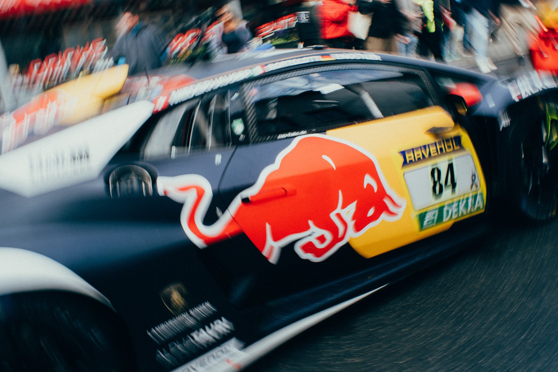 Close-up view of a racing car with a Red Bull logo, the number 84 on the side, and sponsors' decals, in a racing event with spectators in the background.