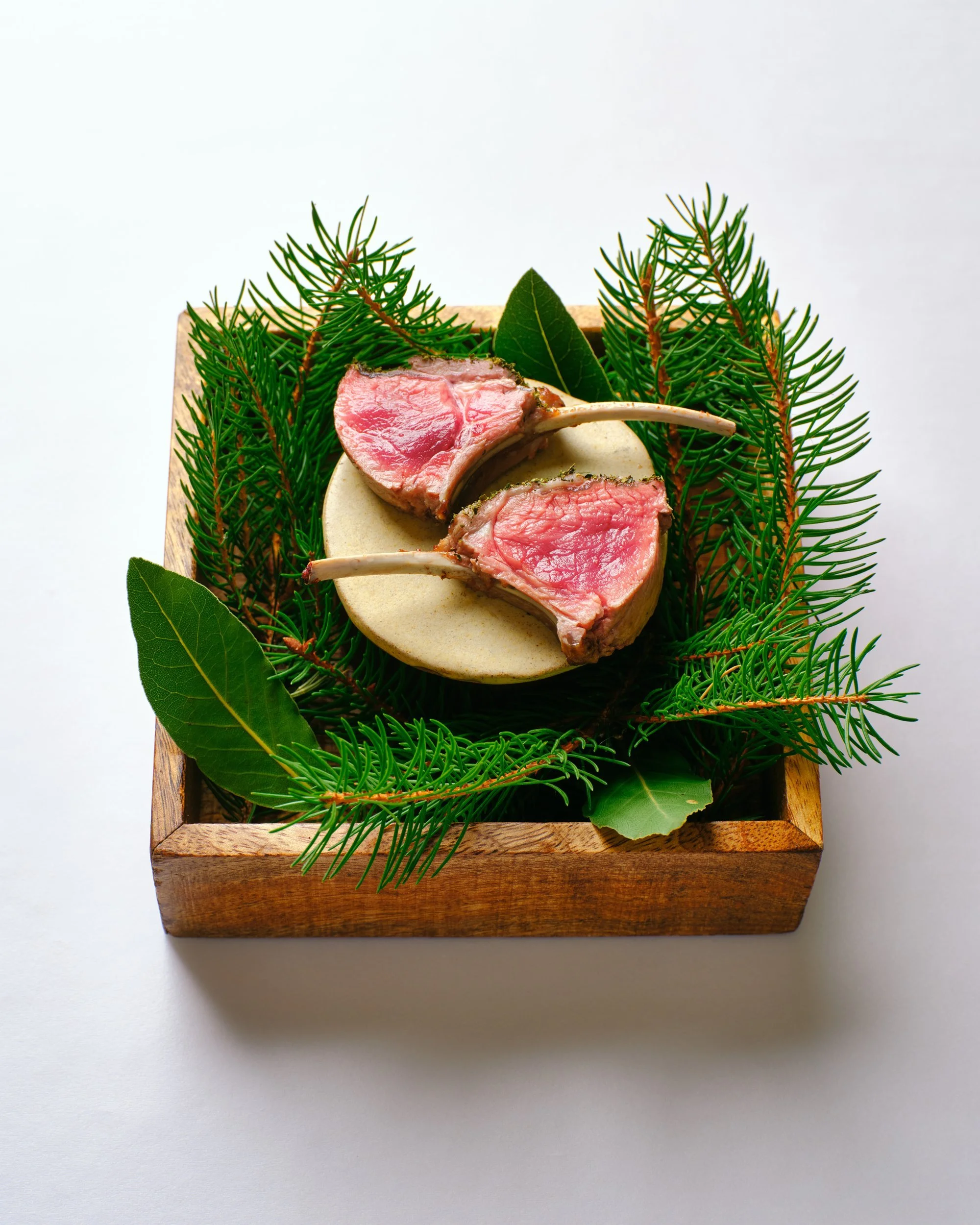 Pink slices of roast lamb on a small round plate, set in a wooden box filled with pine sprigs and bay leaves, on a white background.