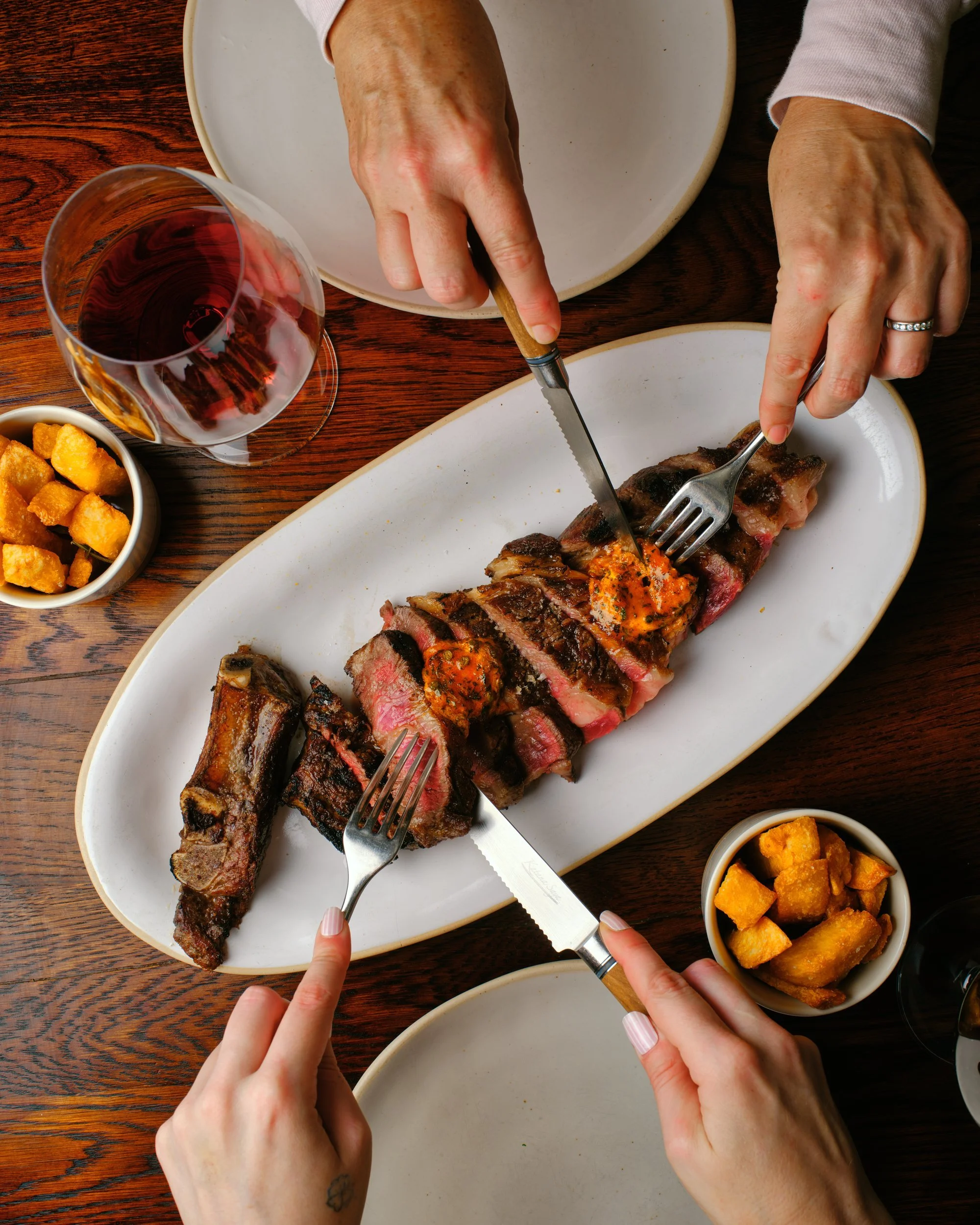 Overhead view of two people slicing into a sharing steak on an oval platter, with sides of crispy potatoes and glasses of red wine on a wooden table.