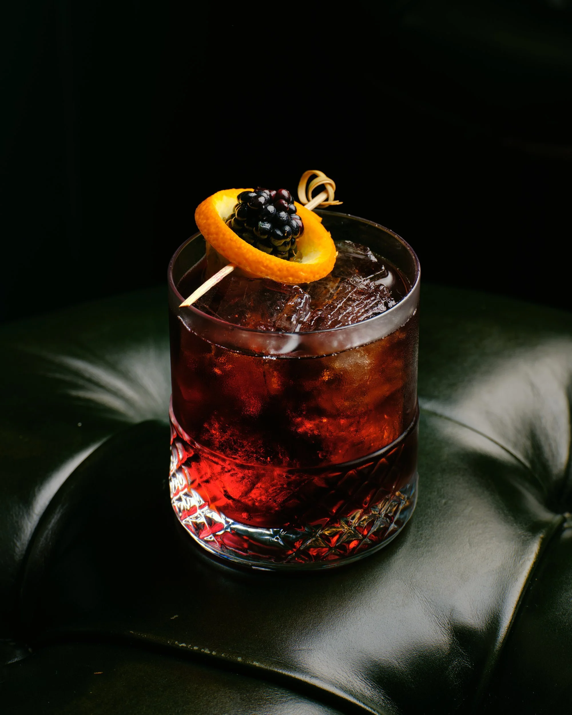 A dark red cocktail in a cut-glass tumbler with an orange twist and a blackberry, against a deep black leather background.