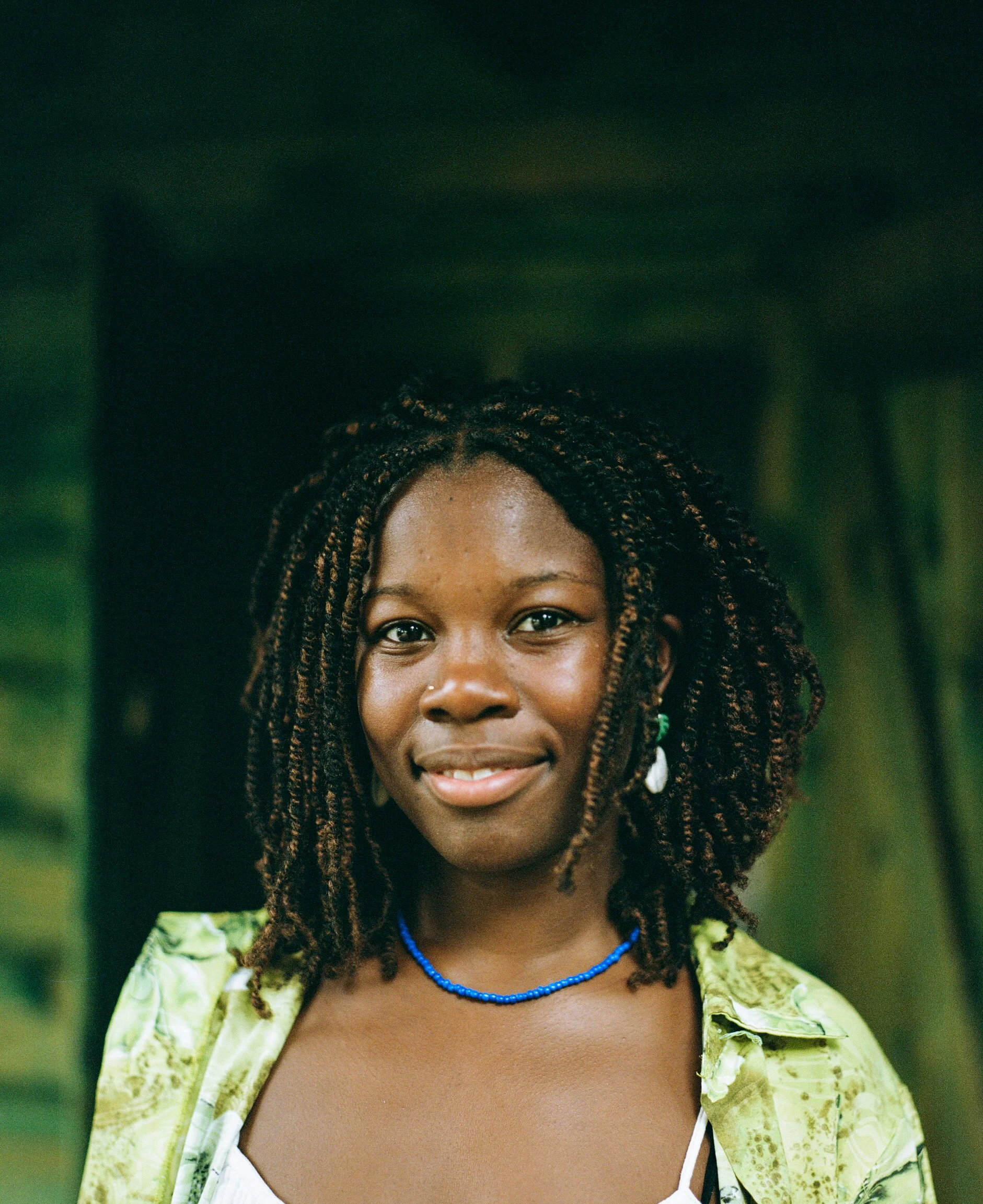 Portrait of a smiling Black woman with dreadlocks, wearing a yellow-green shirt, necklaces, and earrings, standing outdoors with green foliage in the background.