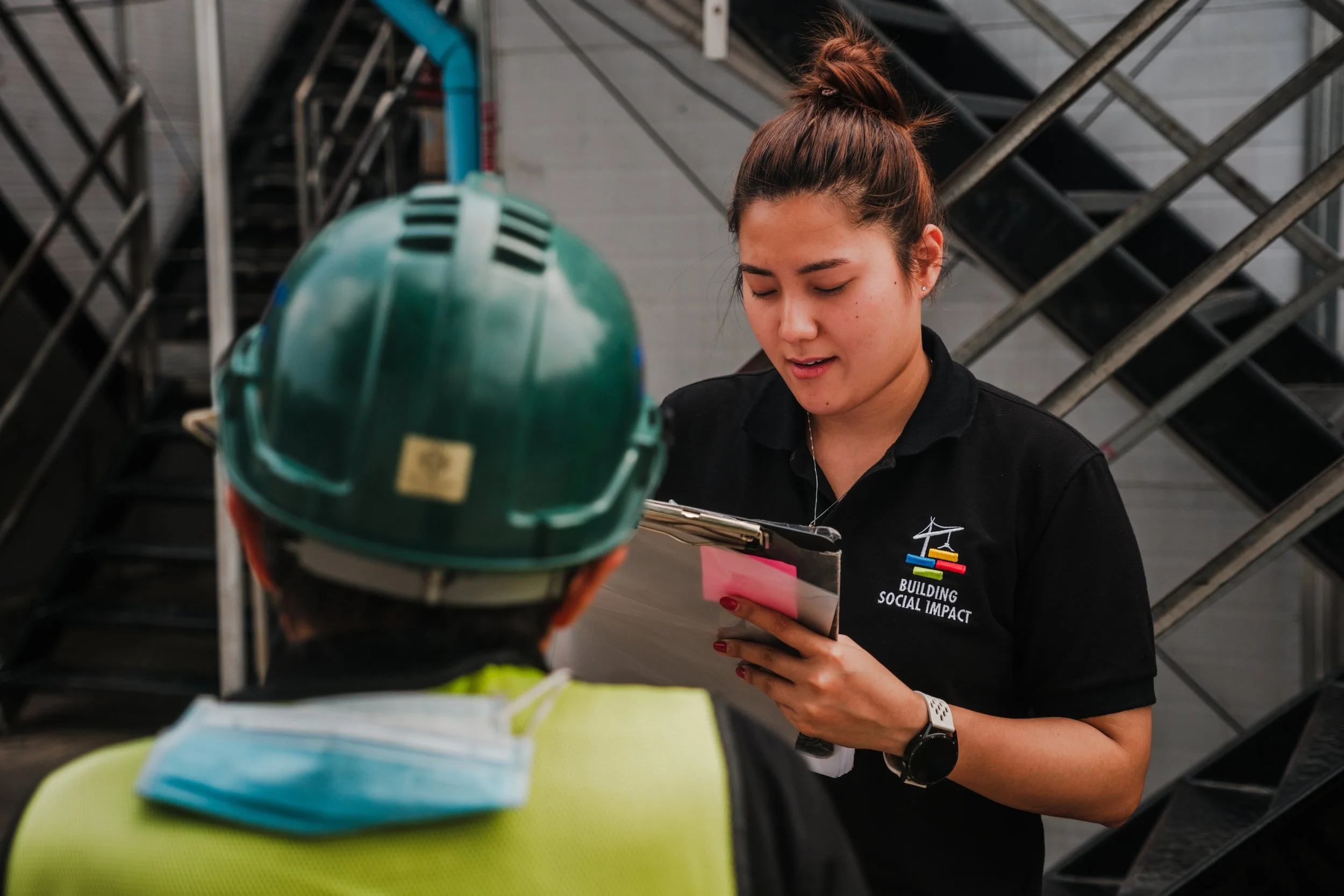 Young woman in a black Polo with the logo 'Building Social Impact' speaking with a person wearing a yellow safety vest and a green hard hat, in an industrial setting.