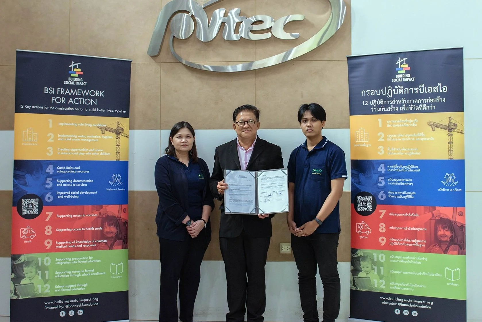 Three people standing in front of two banners and a large 'pVtec' sign, holding a document in an indoor setting.
