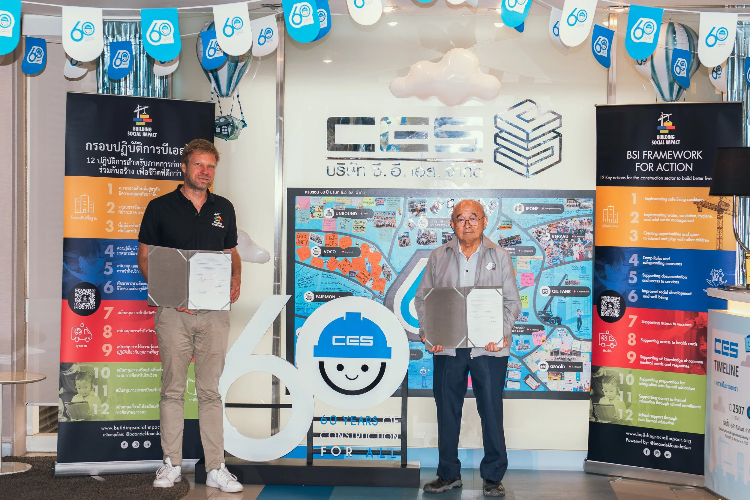 Two men standing inside an office with banners and decorations celebrating 60 years of construction impact, holding documents, with a company logo and a colorful map in the background.