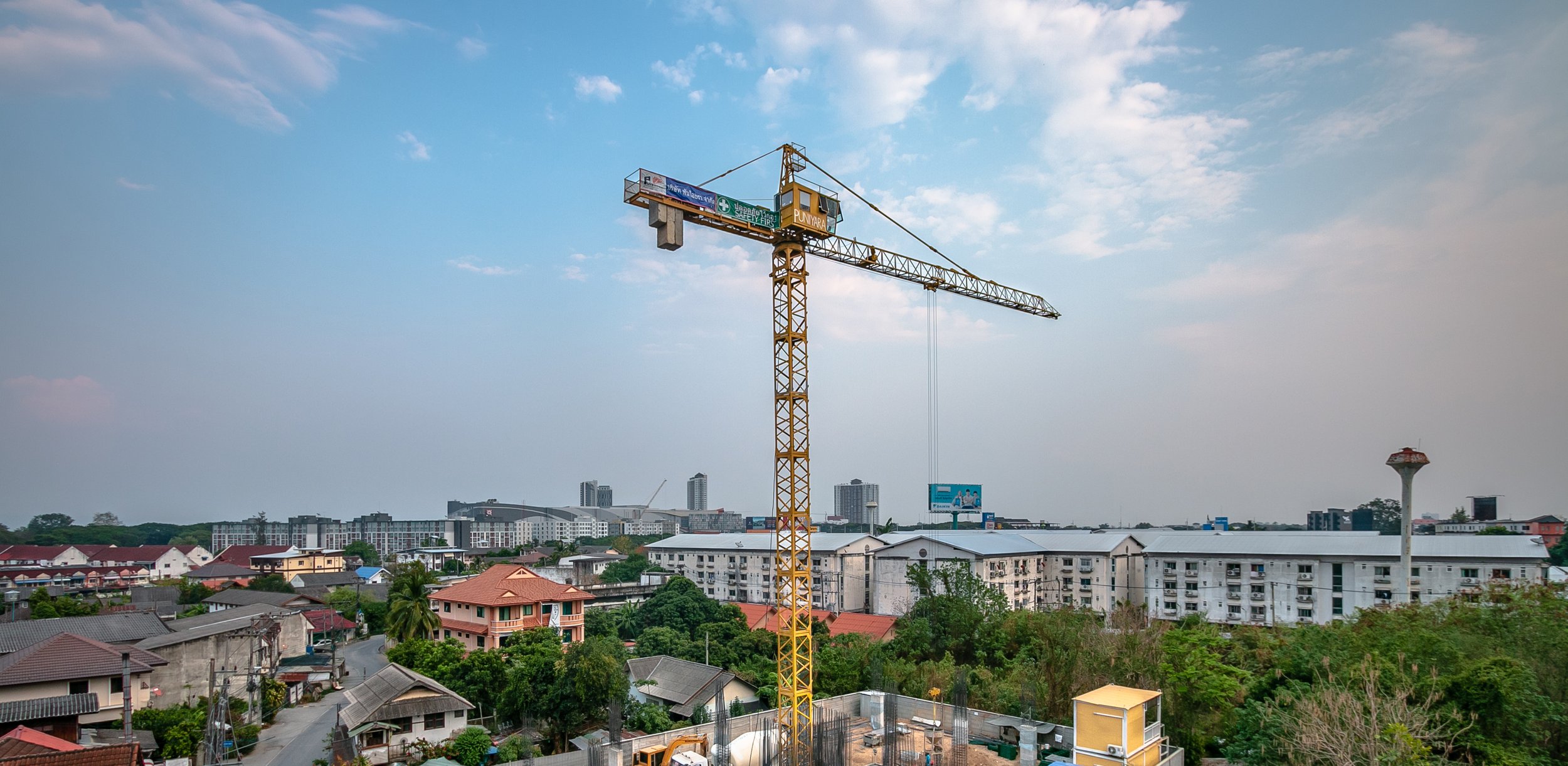 A tall yellow construction crane over a cityscape with residential buildings, trees, and a cloudy sky.