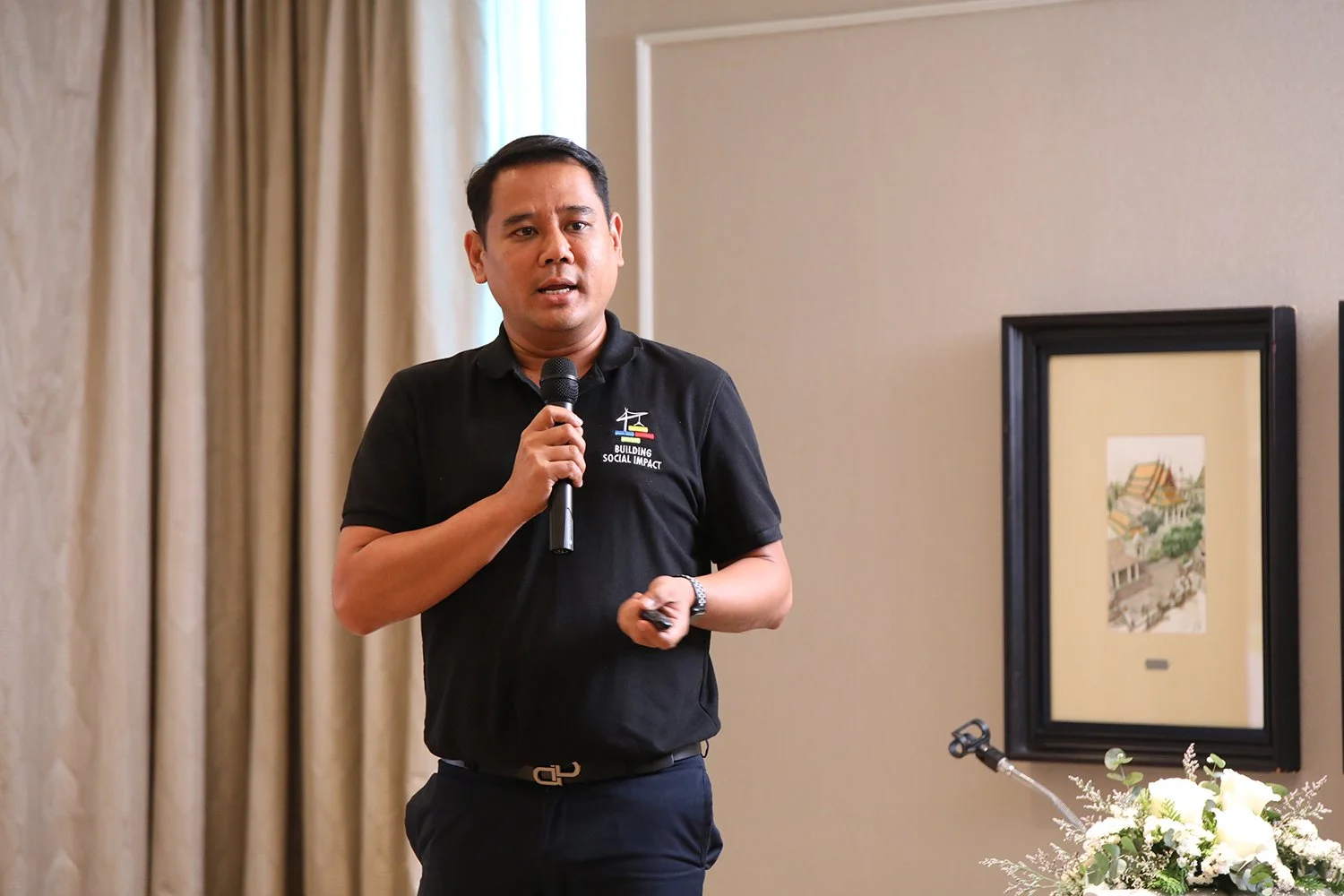 A man holding a microphone and a remote control, giving a presentation in a room with beige wall and framed artwork, wearing a black polo shirt with a logo that reads 'Building Social Impact'.