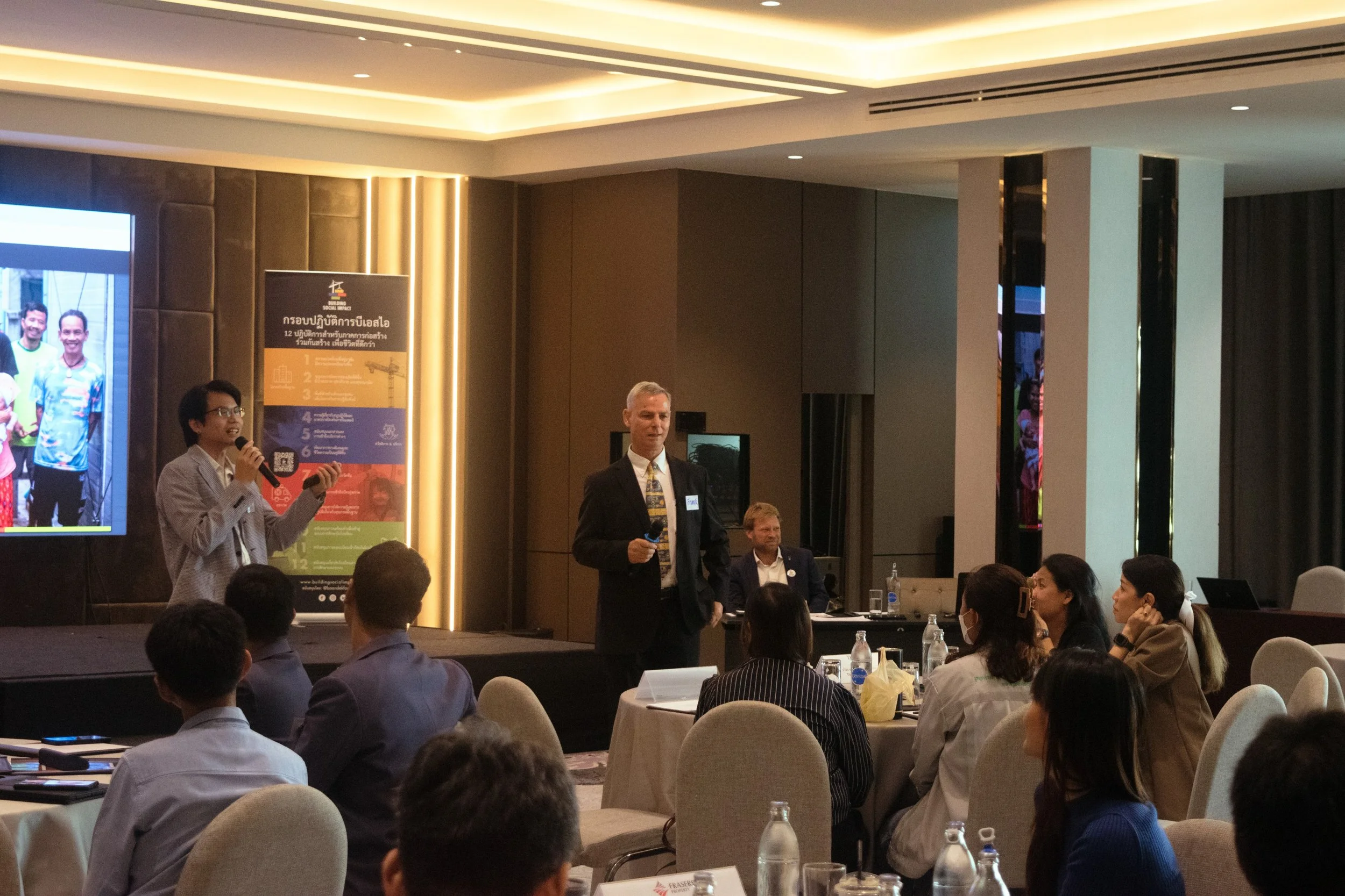 A conference room with a speaker on stage holding a microphone, addressing an audience seated at round tables, with a large screen displaying a presentation and a banner in the background.
