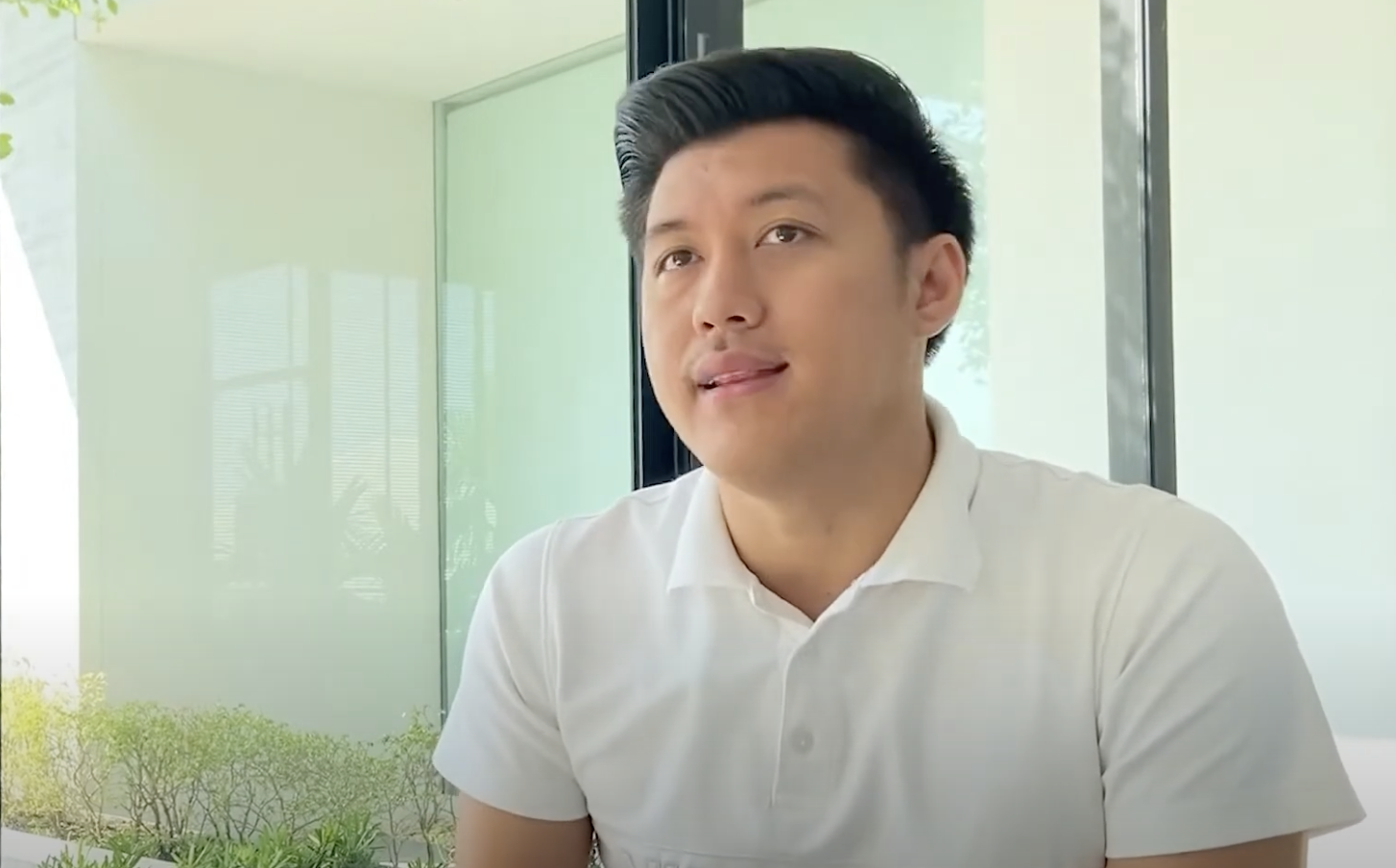 A young man with black hair styled back, wearing a white polo shirt, sitting indoors near a window with a view of greenery outside.