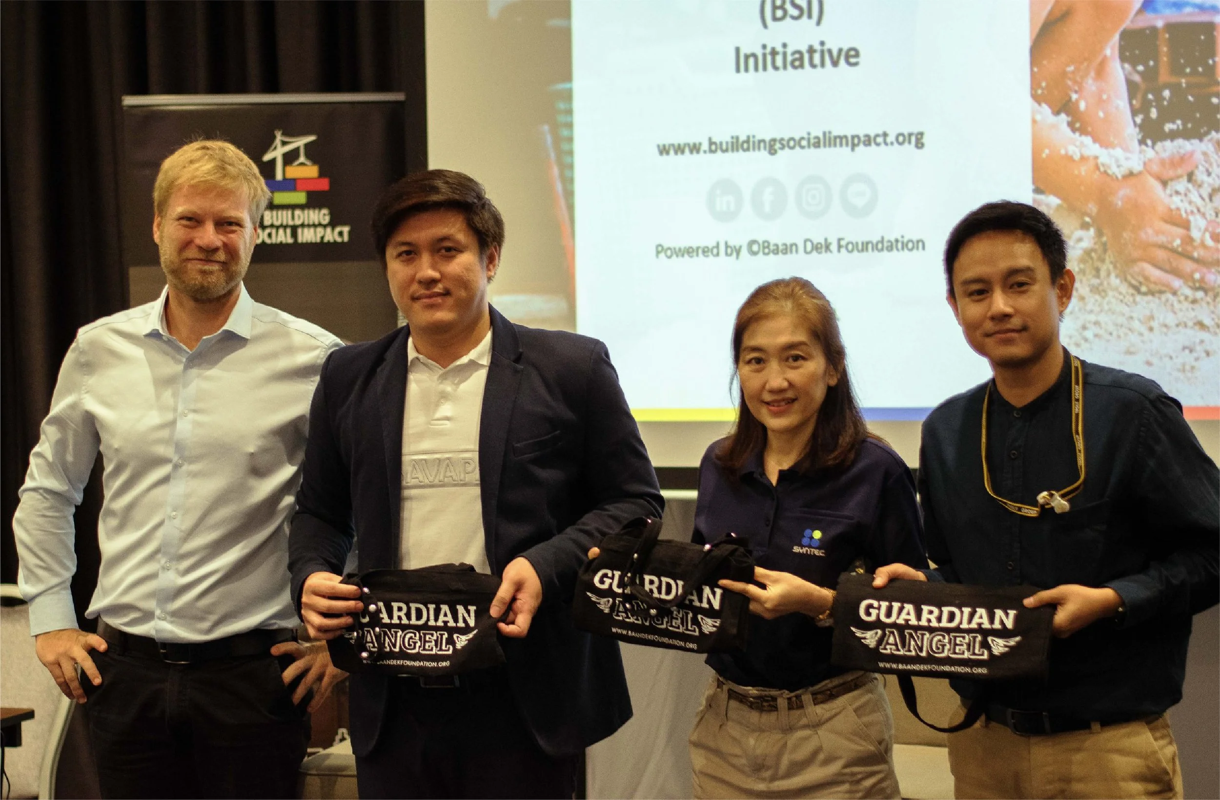Four people standing together at an event, holding black bags with 'Guardian Angel' written on them. A presentation slide and banner are visible in the background.