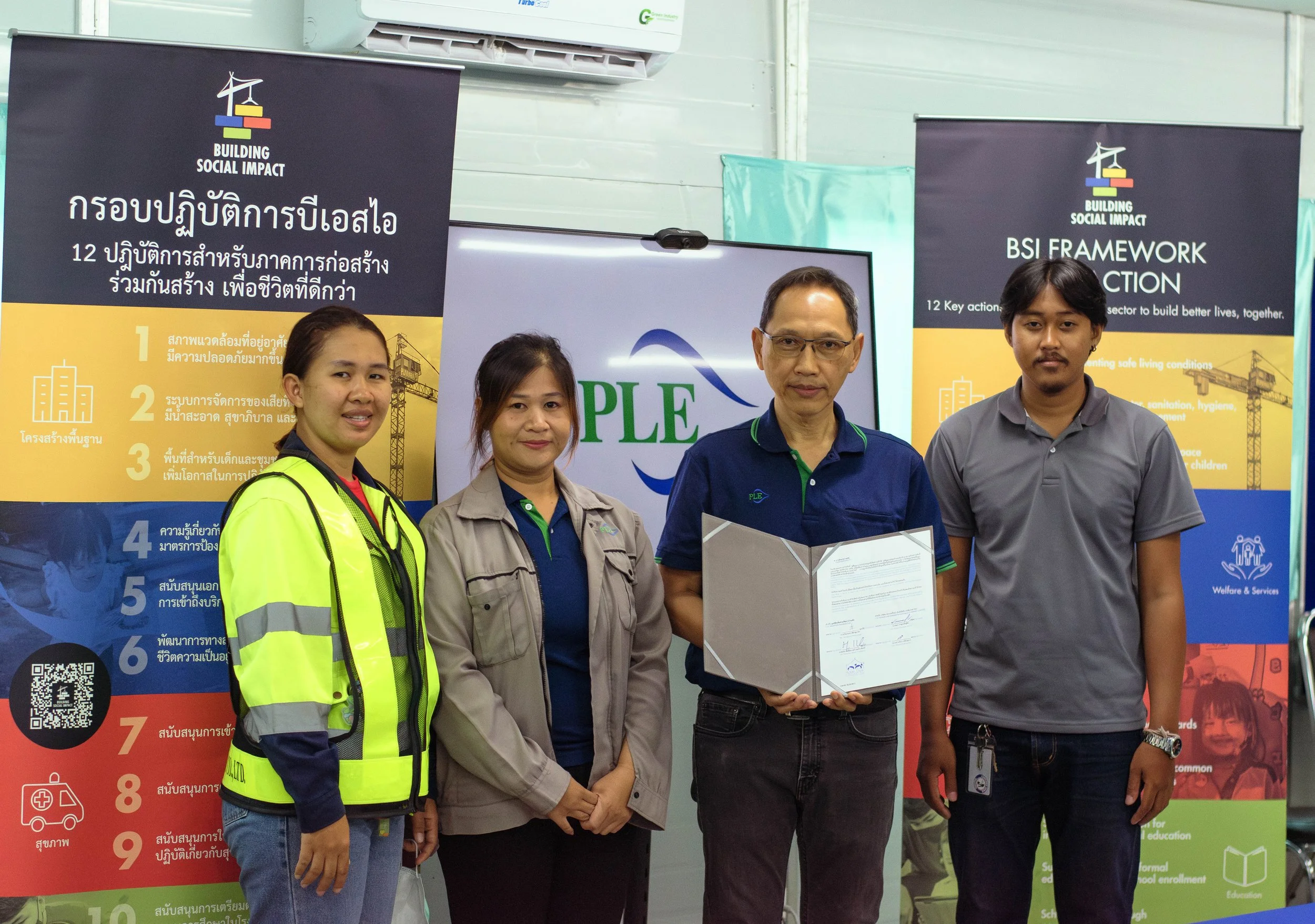 Four people standing in front of banners and a screen at an indoor event, one man holding a certificate, all wearing casual or work attire.