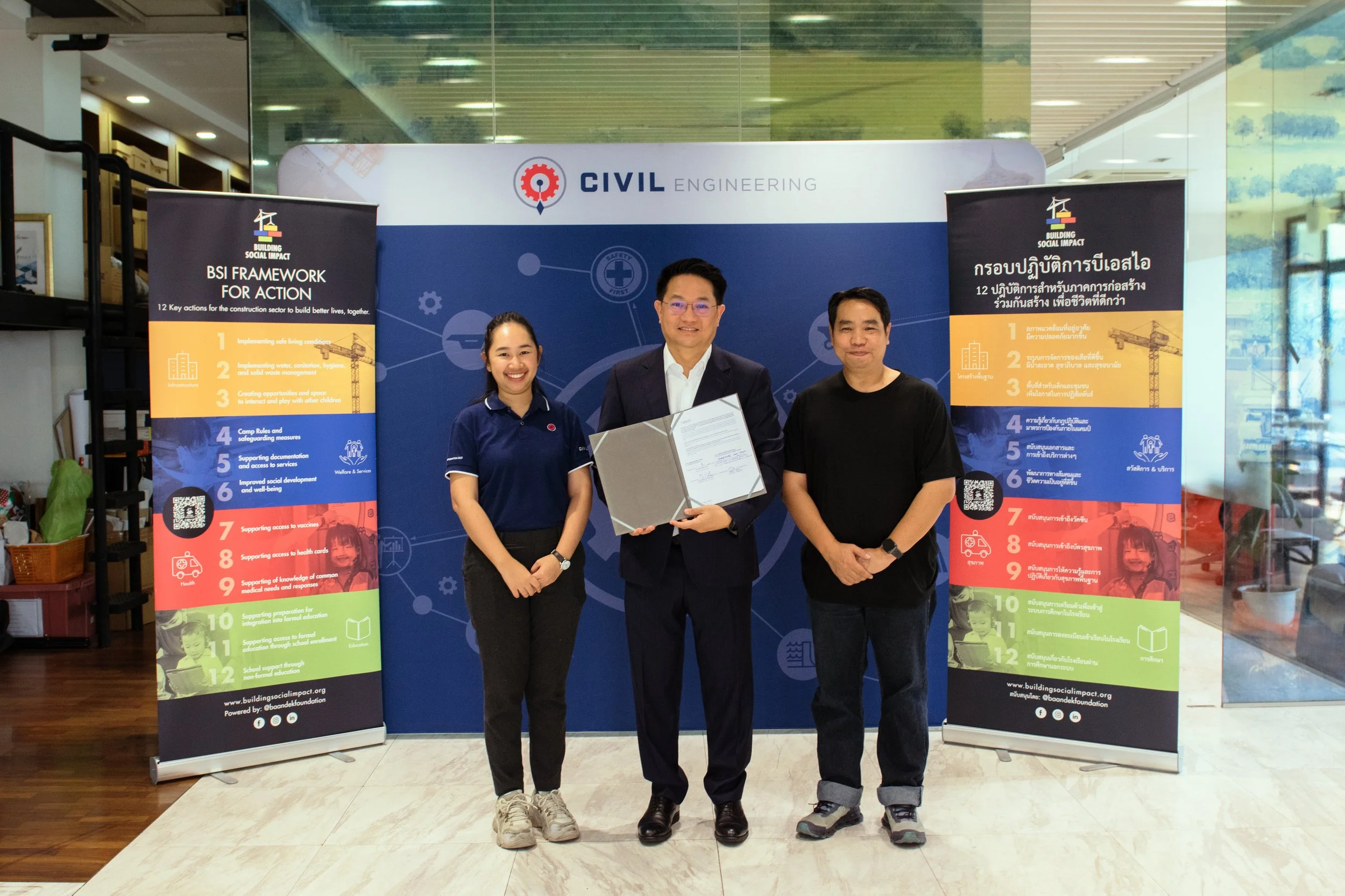 Three people standing in front of a blue backdrop with the logo 'Civil Engineering,' holding a certificate. Two vertical banners on either side display information about the 'Building Social Impact' framework, with text in both English and Thai.