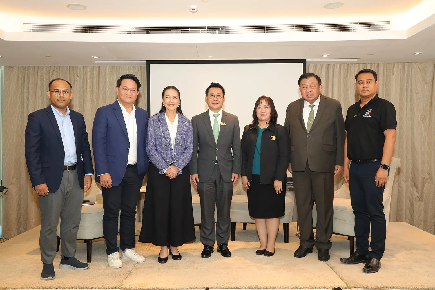 A group of eight professionals standing in a row indoors, dressed in business attire, in front of a classroom or conference room setting with a projection screen.